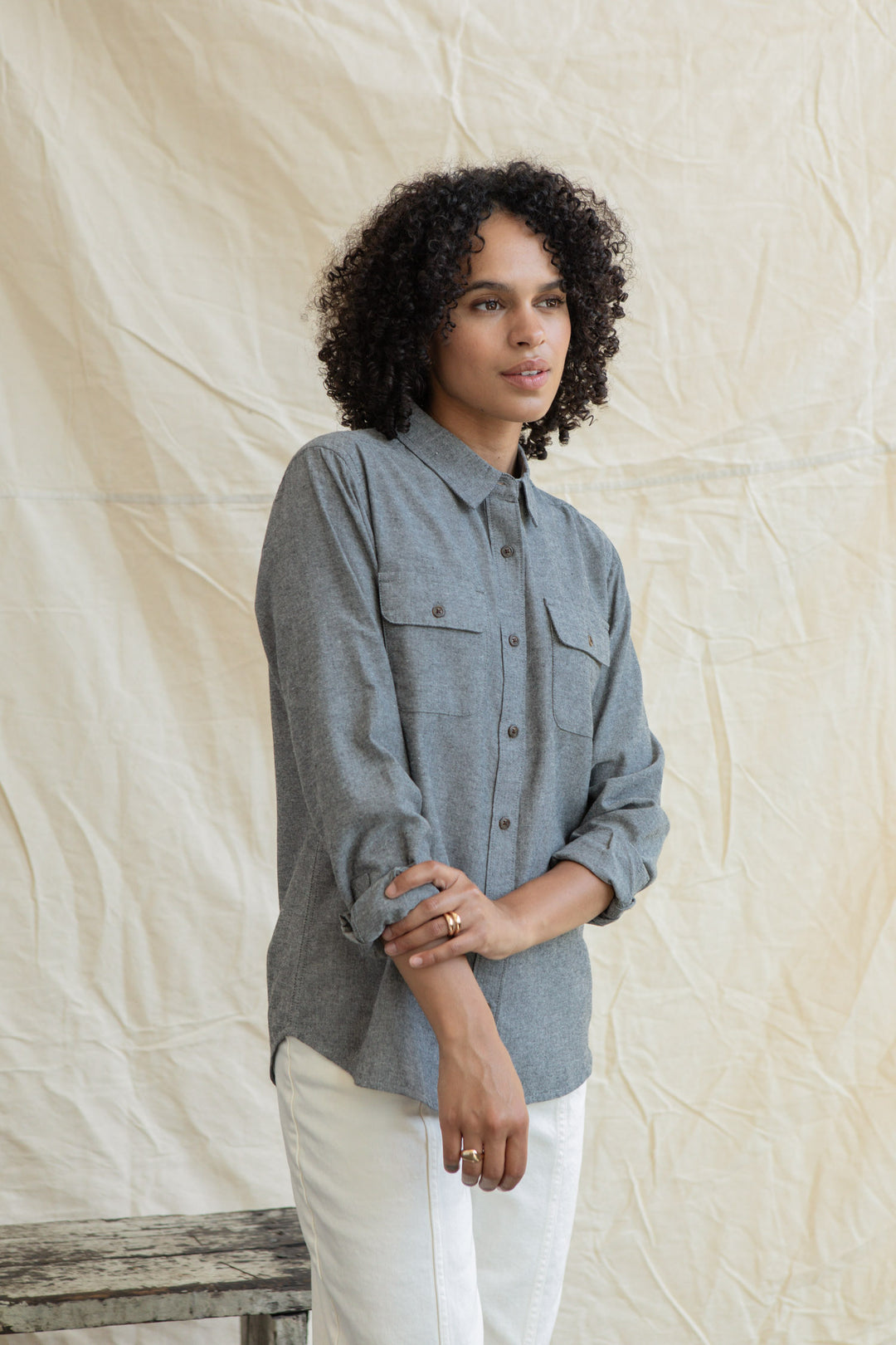 A person with curly hair wears the Bridge & Burn Lenon Shirt in Iron Chambray and white pants, standing before a beige fabric backdrop, holding their left arm with their right hand and looking slightly to the side.