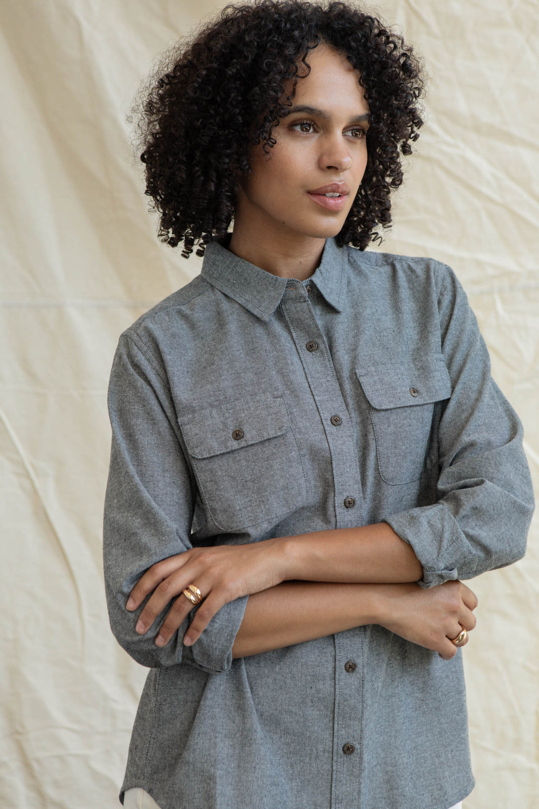 A person with curly hair, arms crossed, wears the Bridge & Burn Lenon Shirt in Iron Chambray with rolled sleeves, standing before a light beige fabric backdrop.
