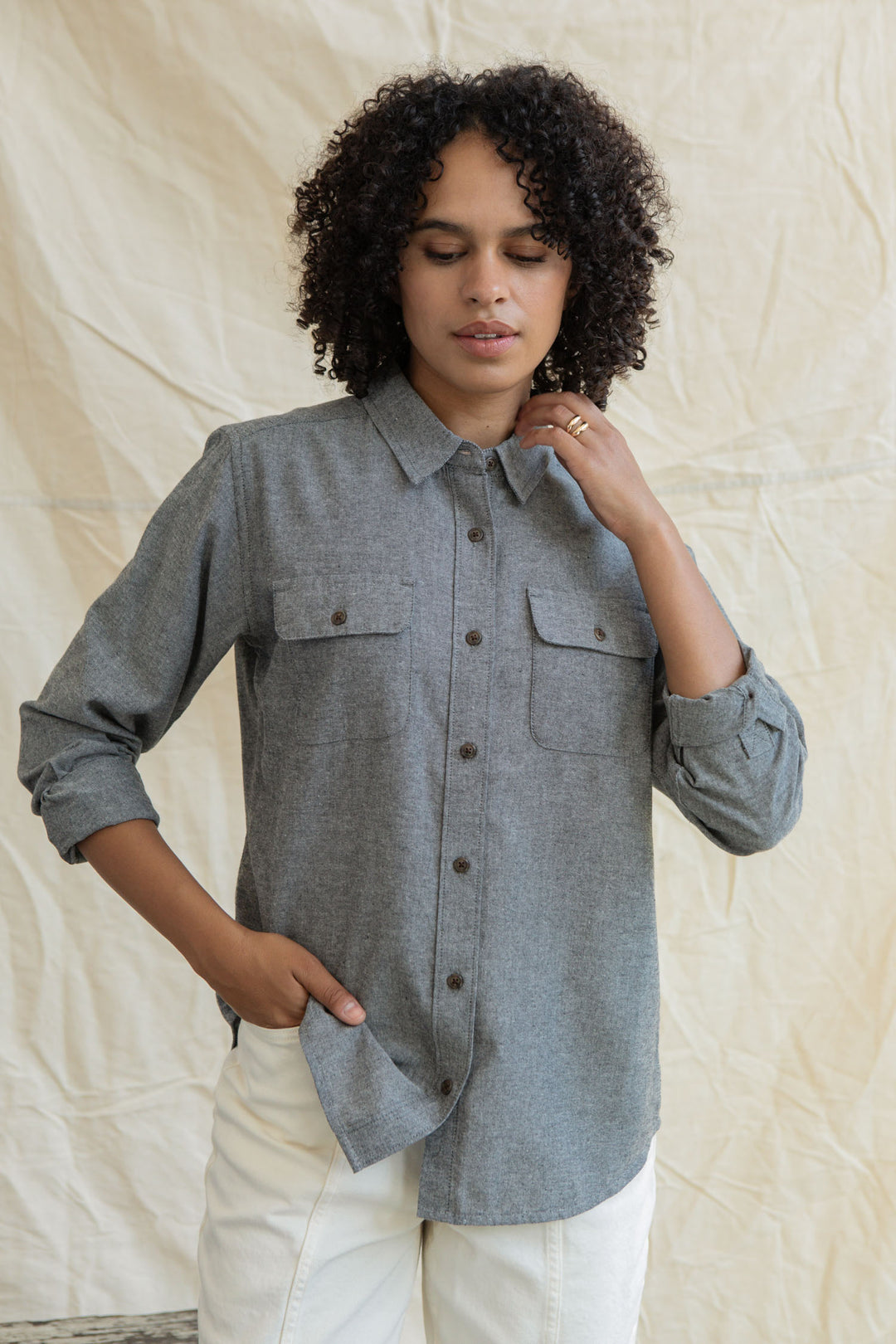 A person with curly hair wears the Bridge & Burn Lenon Shirt in Iron Chambray and white pants, standing before a light beige fabric backdrop, looking down with one hand in their pocket and the other touching their chin.