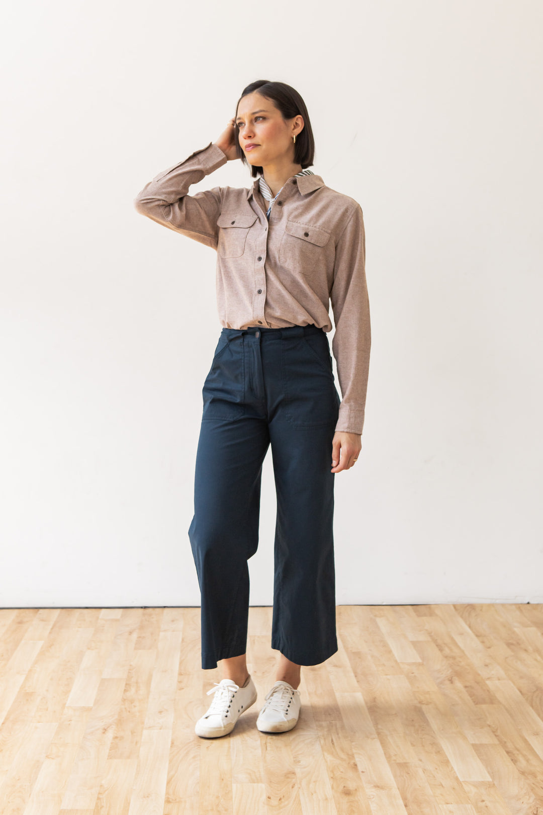 A woman stands indoors on a wooden floor wearing the Bridge & Burn Lenon Shirt in Rust Chambray, navy high-waisted wide-leg pants, and white sneakers, with one hand touching her head as she looks off to the side.