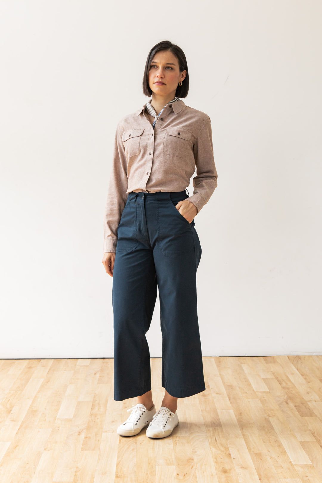 A woman with short dark hair stands indoors on a wooden floor, wearing the Lenon Shirt in Rust Chambray by Bridge & Burn, dark blue high-waisted wide-leg pants, and white sneakers. One hand is in her pocket and she has a neutral expression.