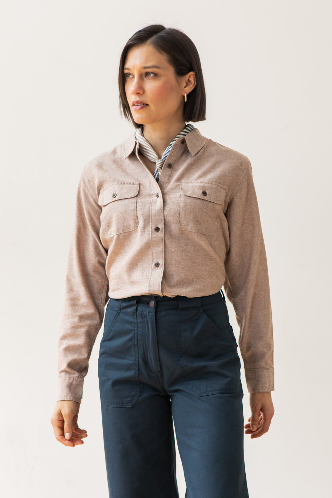 A person with short dark hair wears the Bridge & Burn Lenon Shirt in Rust Chambray over a striped collared shirt and high-waisted navy blue pants, standing against a plain light background, facing slightly to the left.