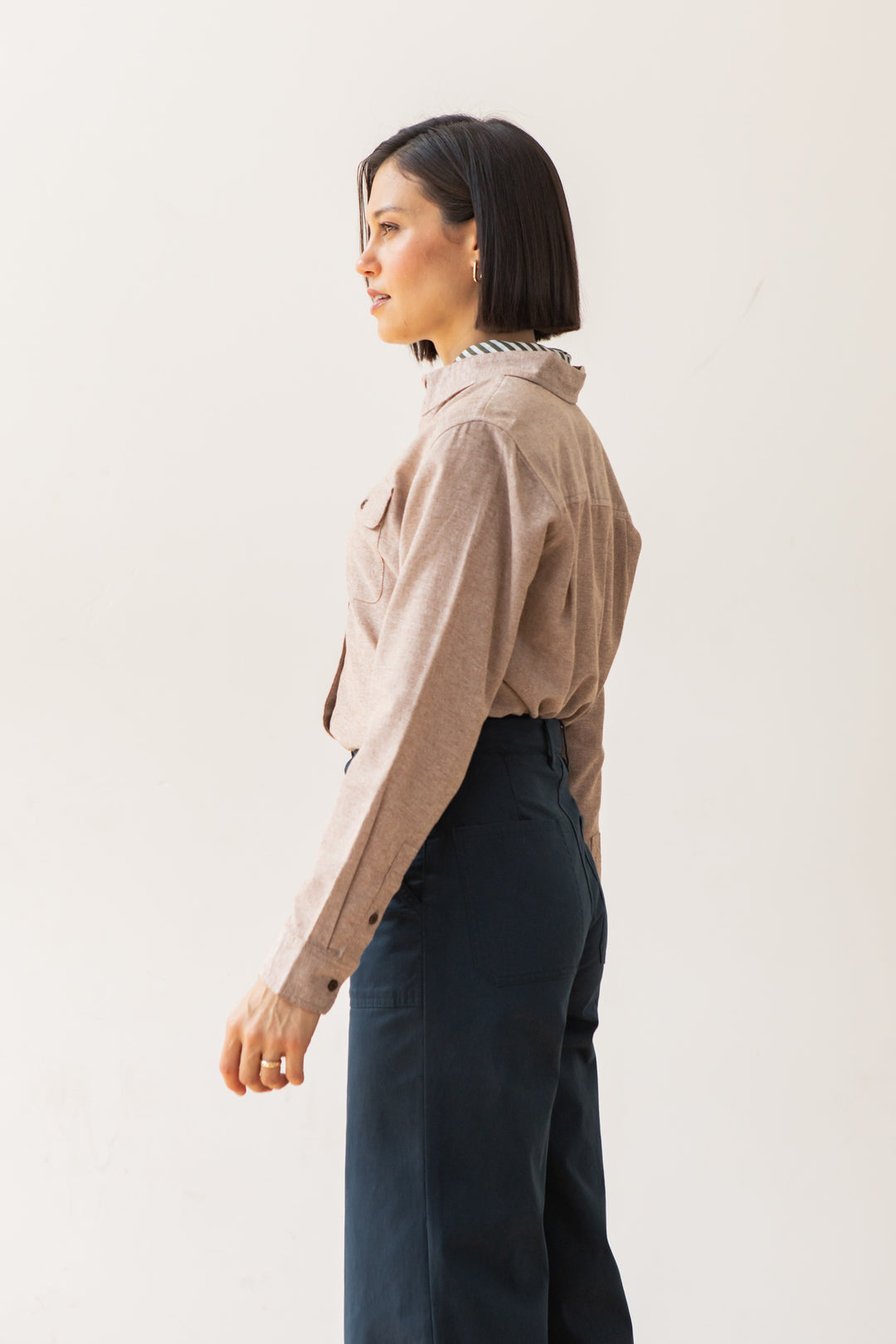 A woman with short dark hair stands in profile against a light background, wearing Bridge & Burn’s Lenon Shirt in Rust Chambray paired with high-waisted dark trousers for an effortlessly refined look.