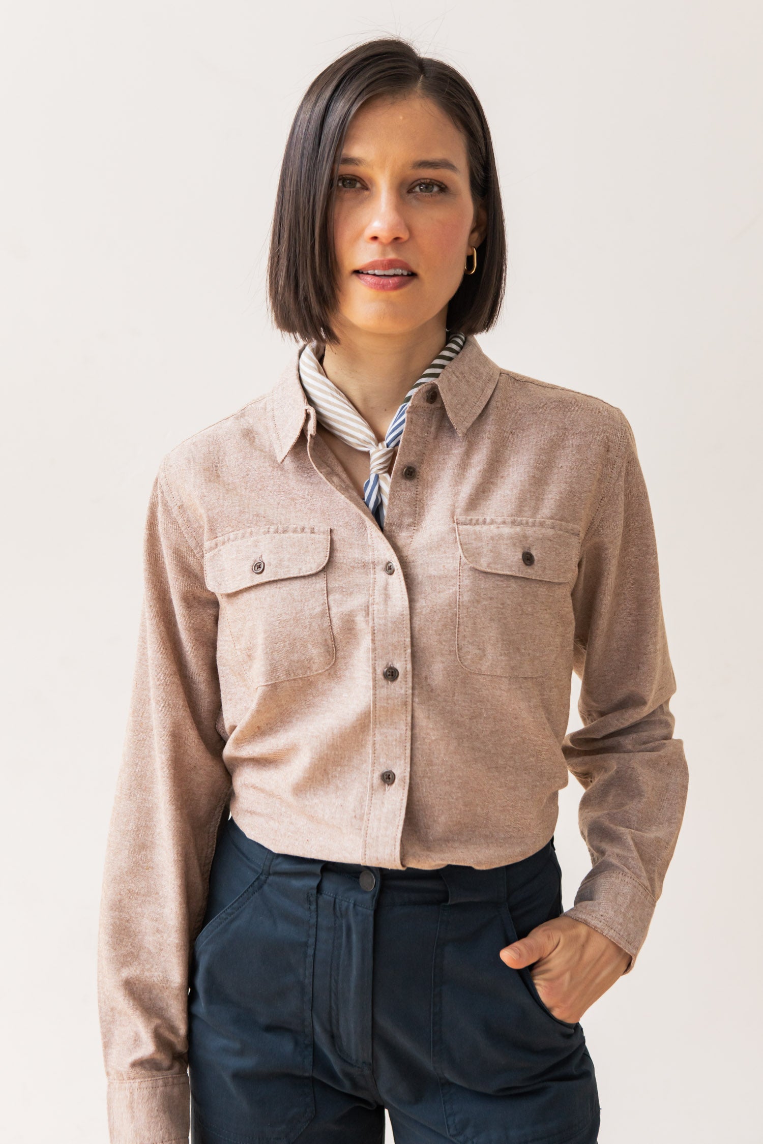 A person with straight, shoulder-length dark hair wears the Bridge & Burn Lenon Shirt in Rust Chambray over a striped collared shirt and dark blue pants, standing against a plain light background.