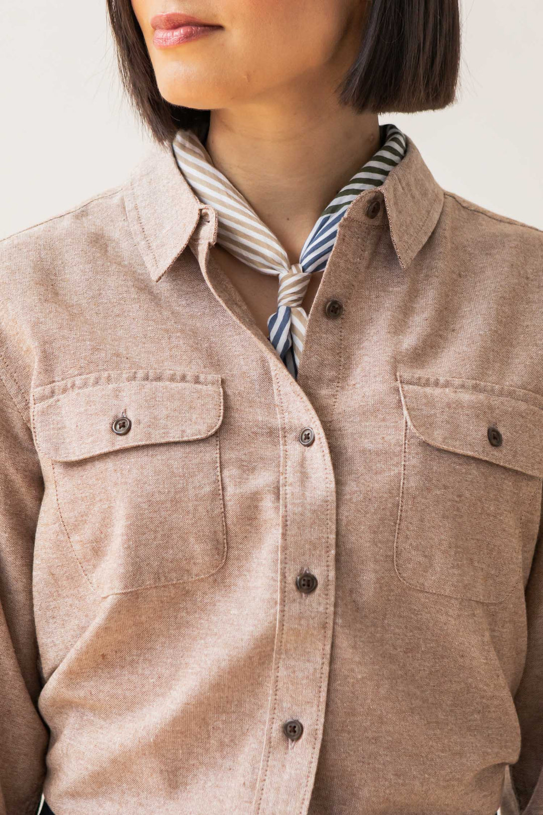 A woman wears the Bridge & Burn Lenon Shirt in Rust Chambray, a relaxed fit style with chest pockets made from recycled cotton, paired with a striped neck scarf. Only her lower face and upper body are visible.