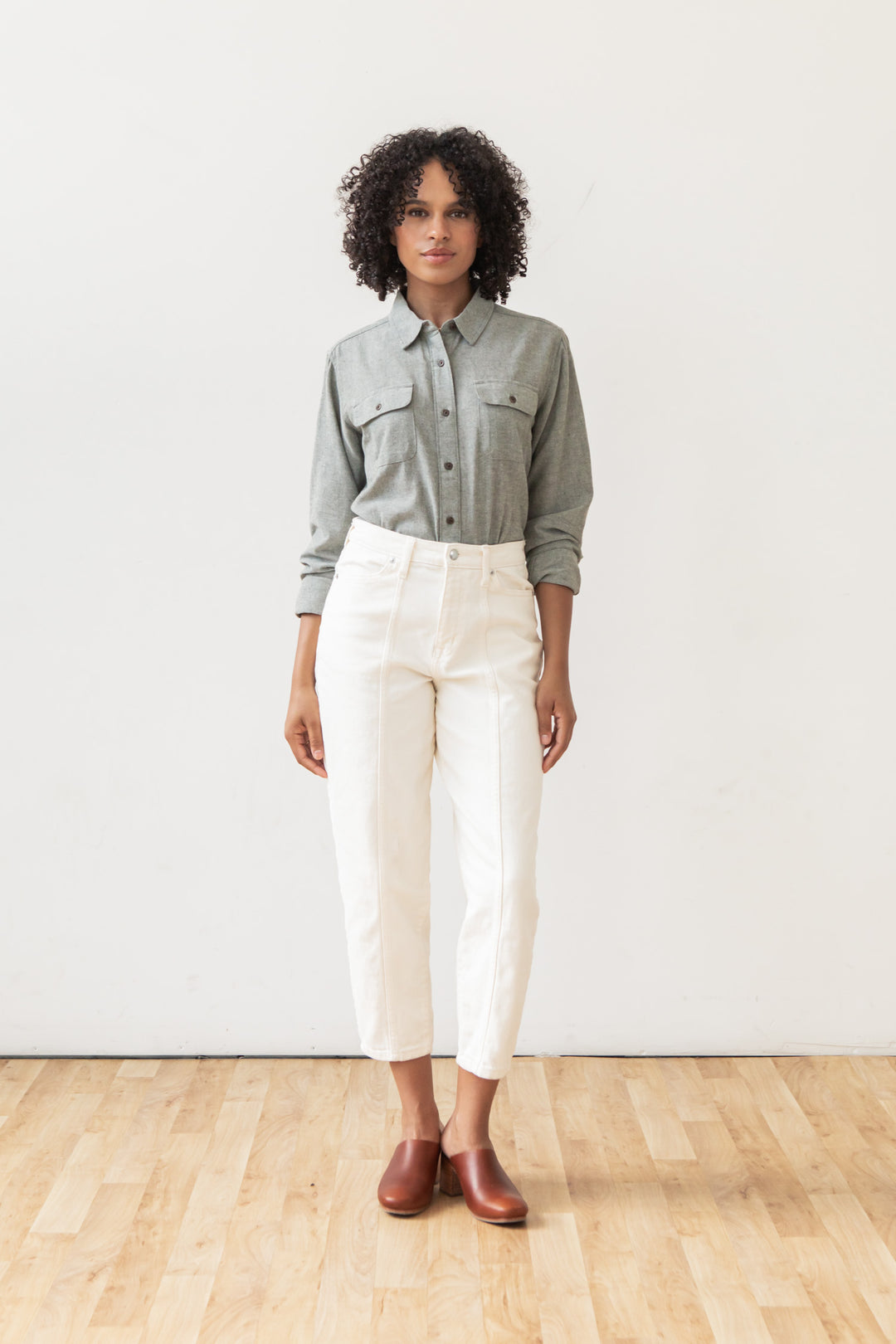 A person with curly hair stands against a white wall, wearing the Bridge & Burn Lenon Shirt in Sage Chambray, white high-waisted pants, and brown slip-on shoes on a wooden floor.
