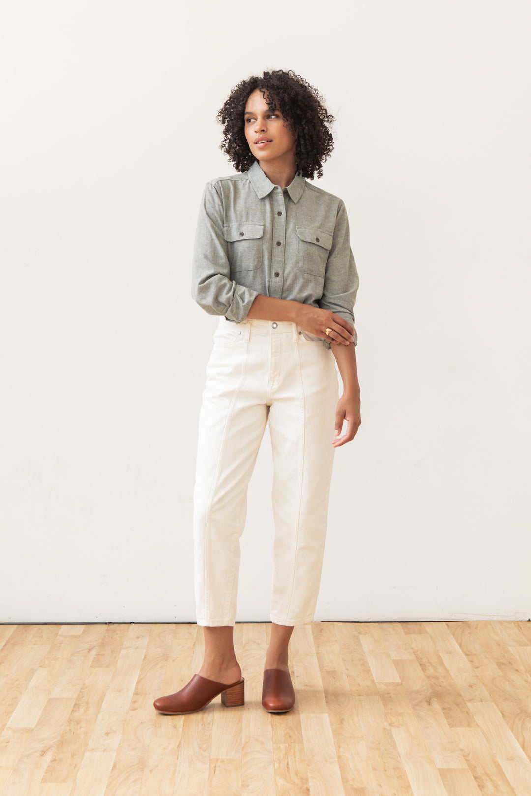 A woman with curly hair stands on a wooden floor against a white wall, wearing the Bridge & Burn Lenon Shirt in Sage Chambray, paired with white high-waisted pants and brown clogs. She looks slightly to the side.