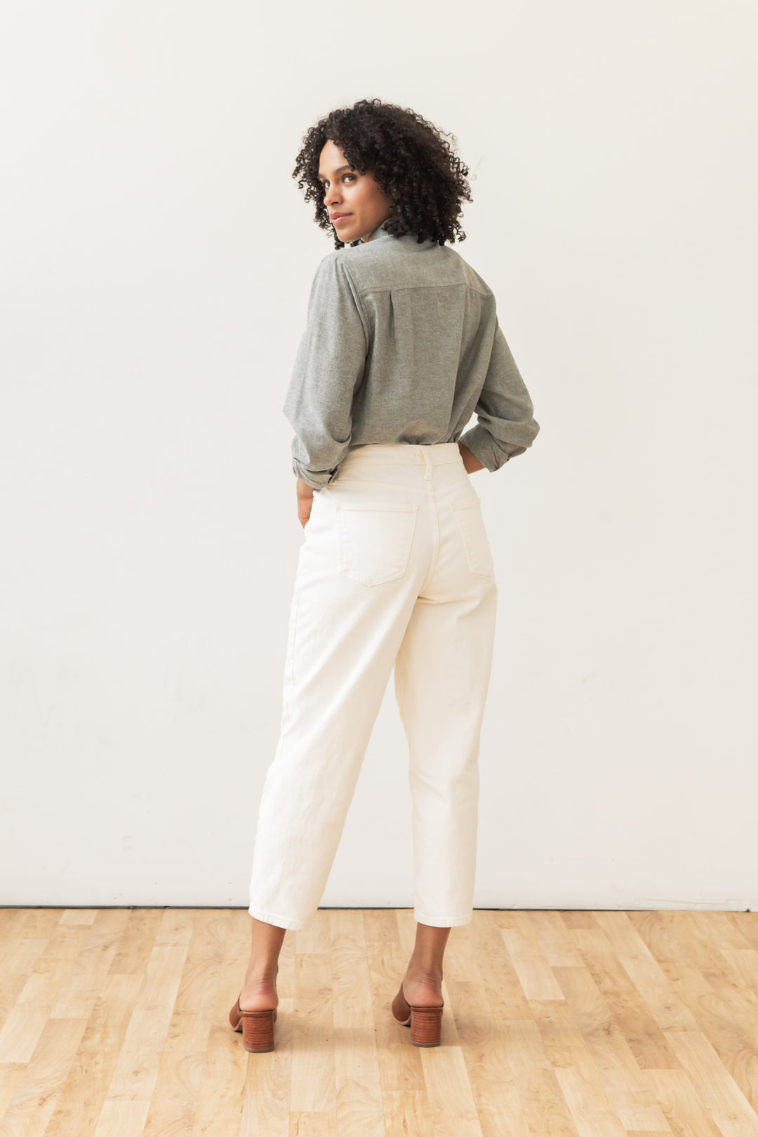 A person with curly hair stands on a wooden floor, facing away and looking over their shoulder, wearing the Bridge & Burn Lenon Shirt in Sage Chambray paired with high-waisted white pants and brown heeled shoes against a white backdrop.