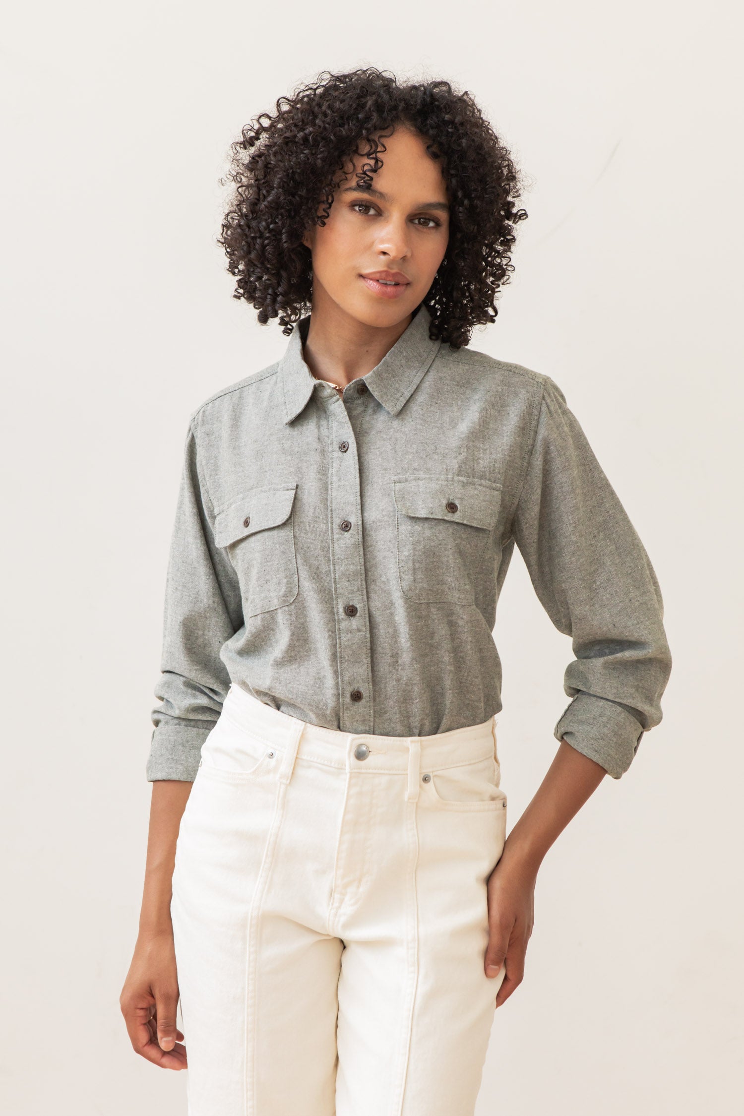 A person with curly hair wears the Lenon Shirt in Sage Chambray by Bridge & Burn and white high-waisted pants, standing confidently against a plain light background.