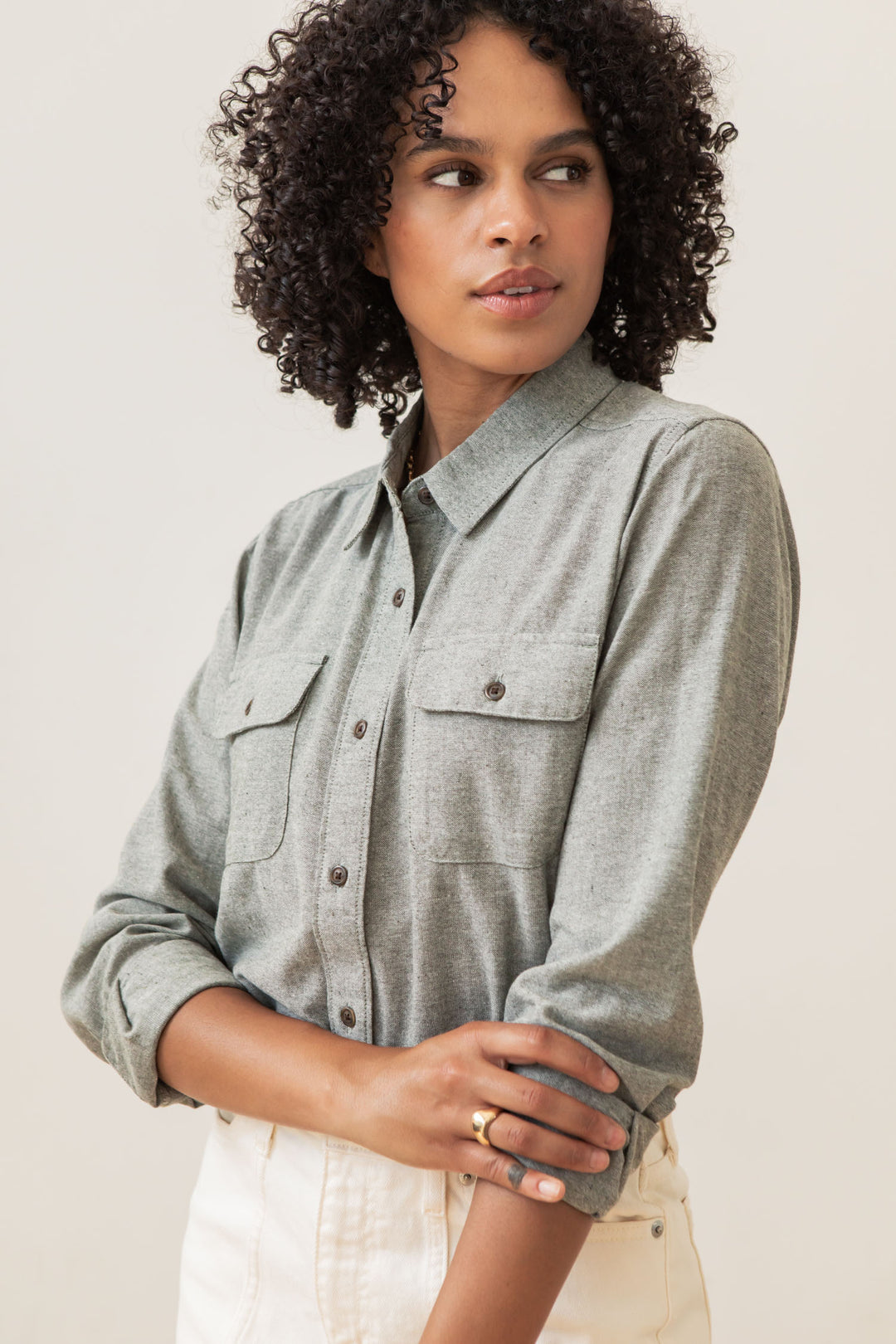 A woman with curly hair wears the Bridge & Burn Lenon Shirt in Sage Chambray with white pants, standing against a plain background and looking to the side with her arms gently crossed.