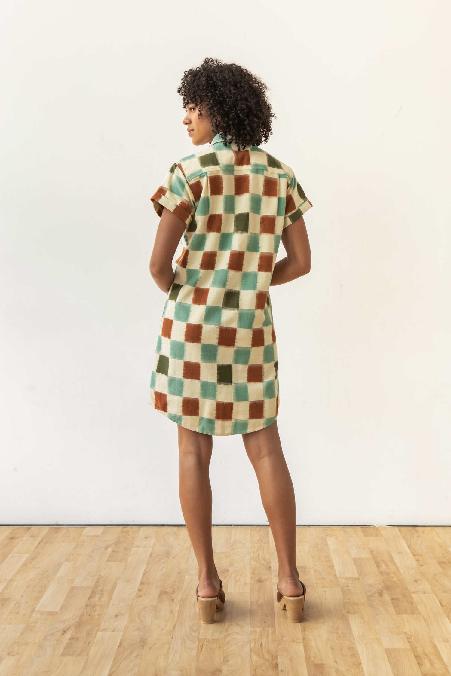 A woman with curly hair stands on a wooden floor, facing away in the Bridge & Burn Loren Shirt Dress / Mod Checker—a green, brown, and cream 100% cotton ikat button-front shirt dress—paired with heeled sandals against a plain light wall.