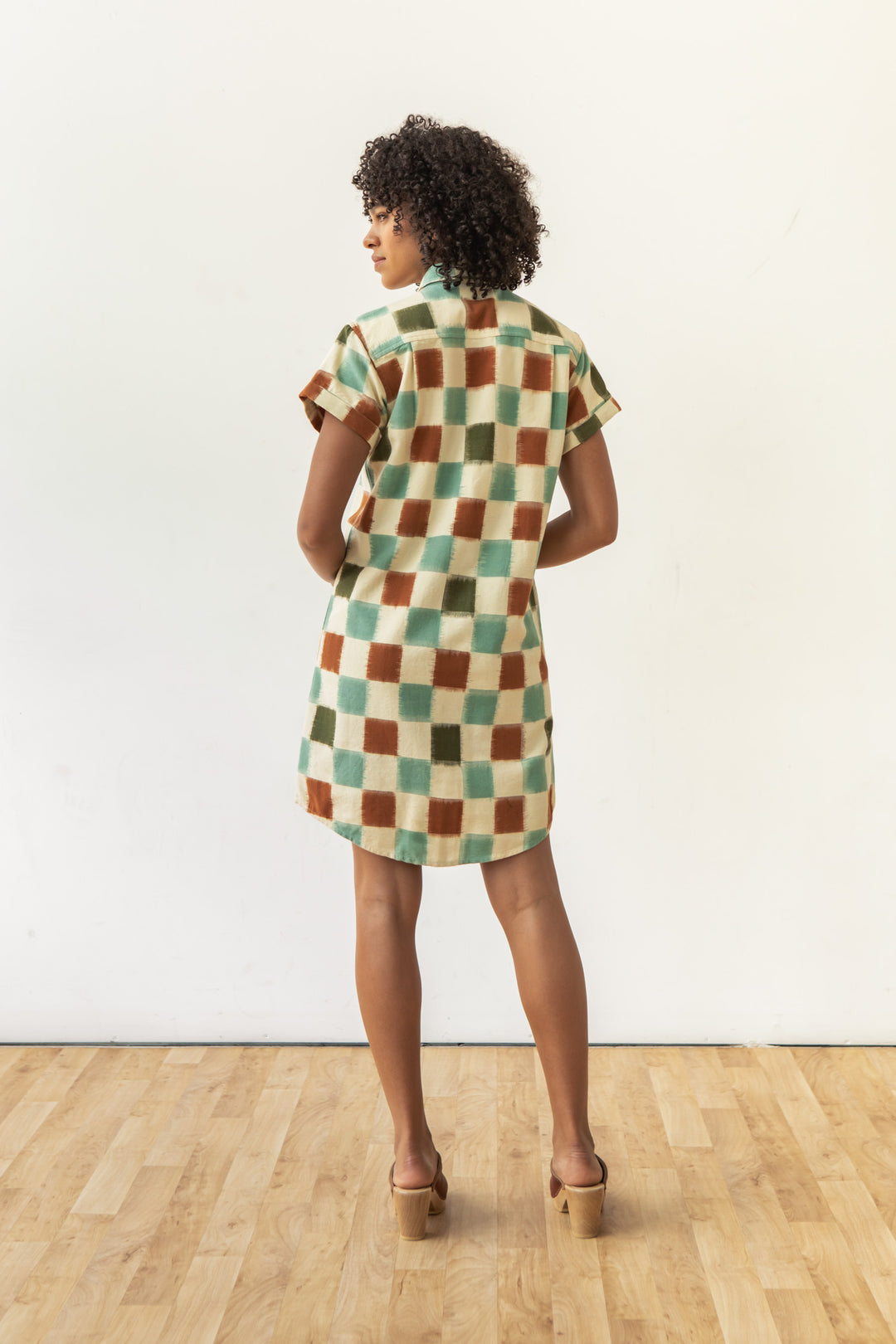 A woman with curly hair stands on a wooden floor, facing away in the Bridge & Burn Loren Shirt Dress / Mod Checker—a green, brown, and cream 100% cotton ikat button-front shirt dress—paired with heeled sandals against a plain light wall.