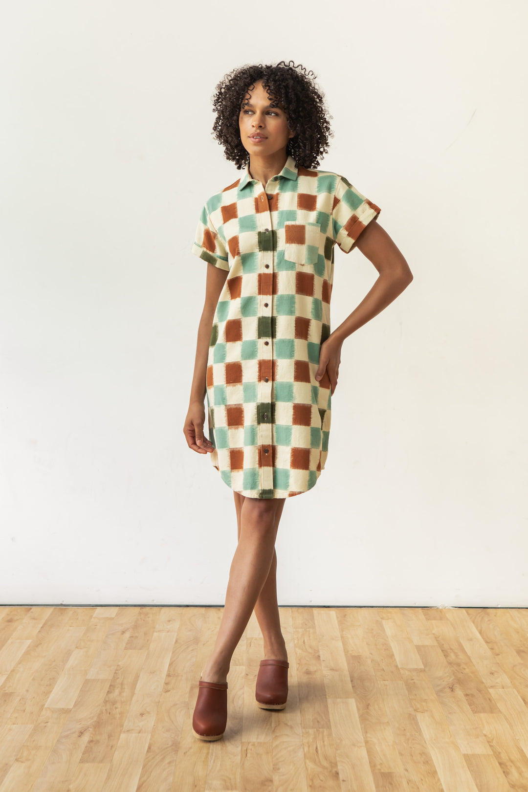 A person with curly hair wears the Bridge & Burn Loren Shirt Dress in Mod Checker— a short-sleeved, 100% cotton button-front dress with a green, rust, and cream check pattern—standing on a wooden floor against a plain white wall.