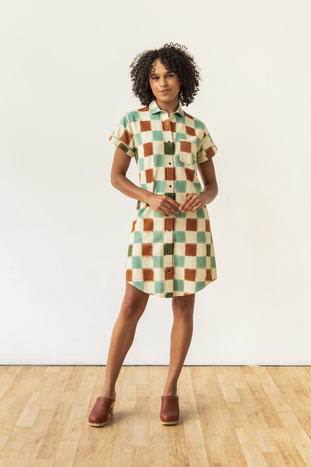A woman with curly hair stands on a wooden floor wearing the Bridge & Burn Loren Shirt Dress in Mod Checker, featuring a green, orange, and cream check pattern. She pairs it with brown shoes and poses against a plain white wall.
