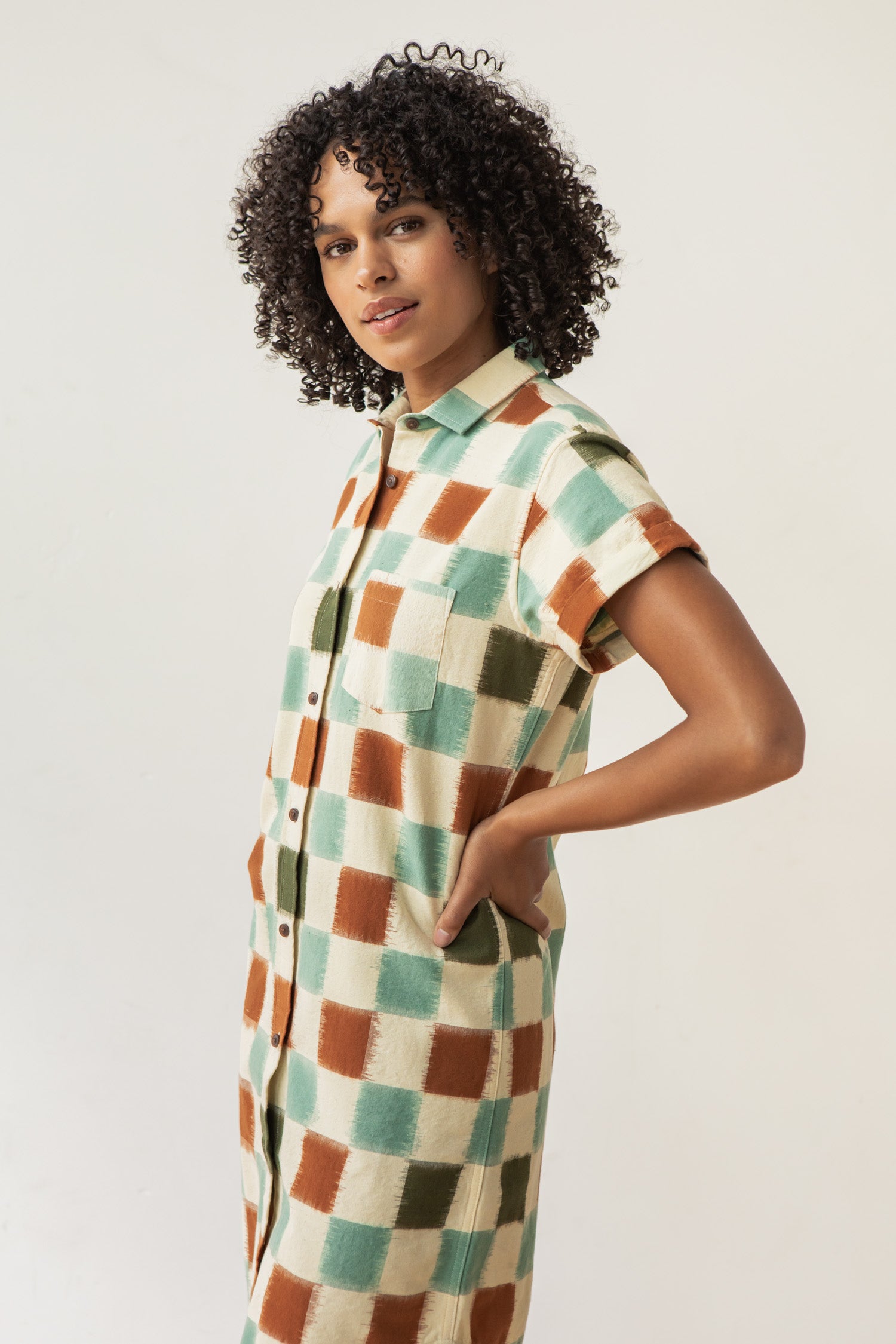 A woman with curly hair stands sideways, hands in pockets, wearing the Bridge & Burn Loren Shirt Dress in Mod Checker—100% cotton with short sleeves, button front, and a bold beige, green, and rust checkered pattern.
