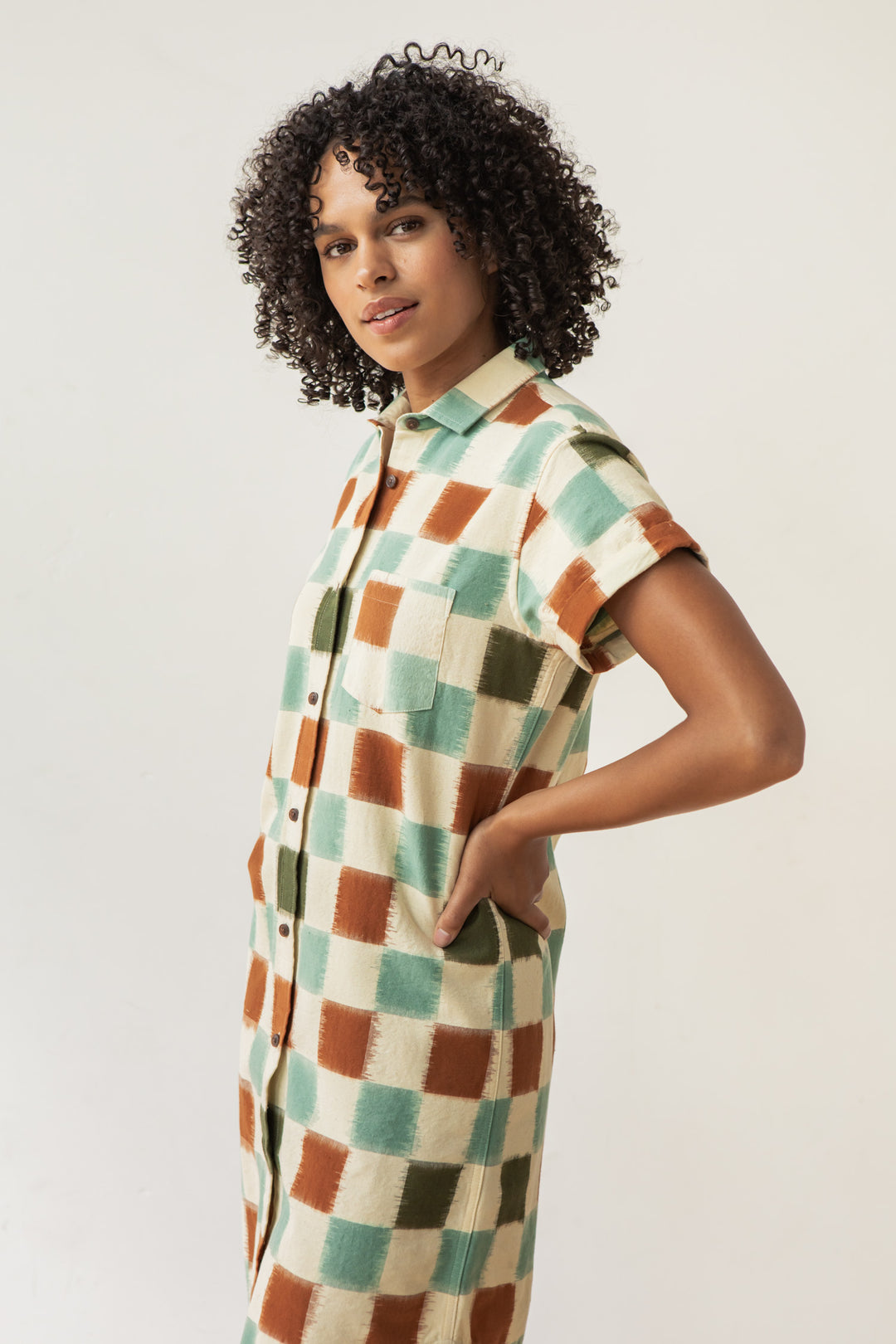 A woman with curly hair stands sideways, hands in pockets, wearing the Bridge & Burn Loren Shirt Dress in Mod Checker—100% cotton with short sleeves, button front, and a bold beige, green, and rust checkered pattern.