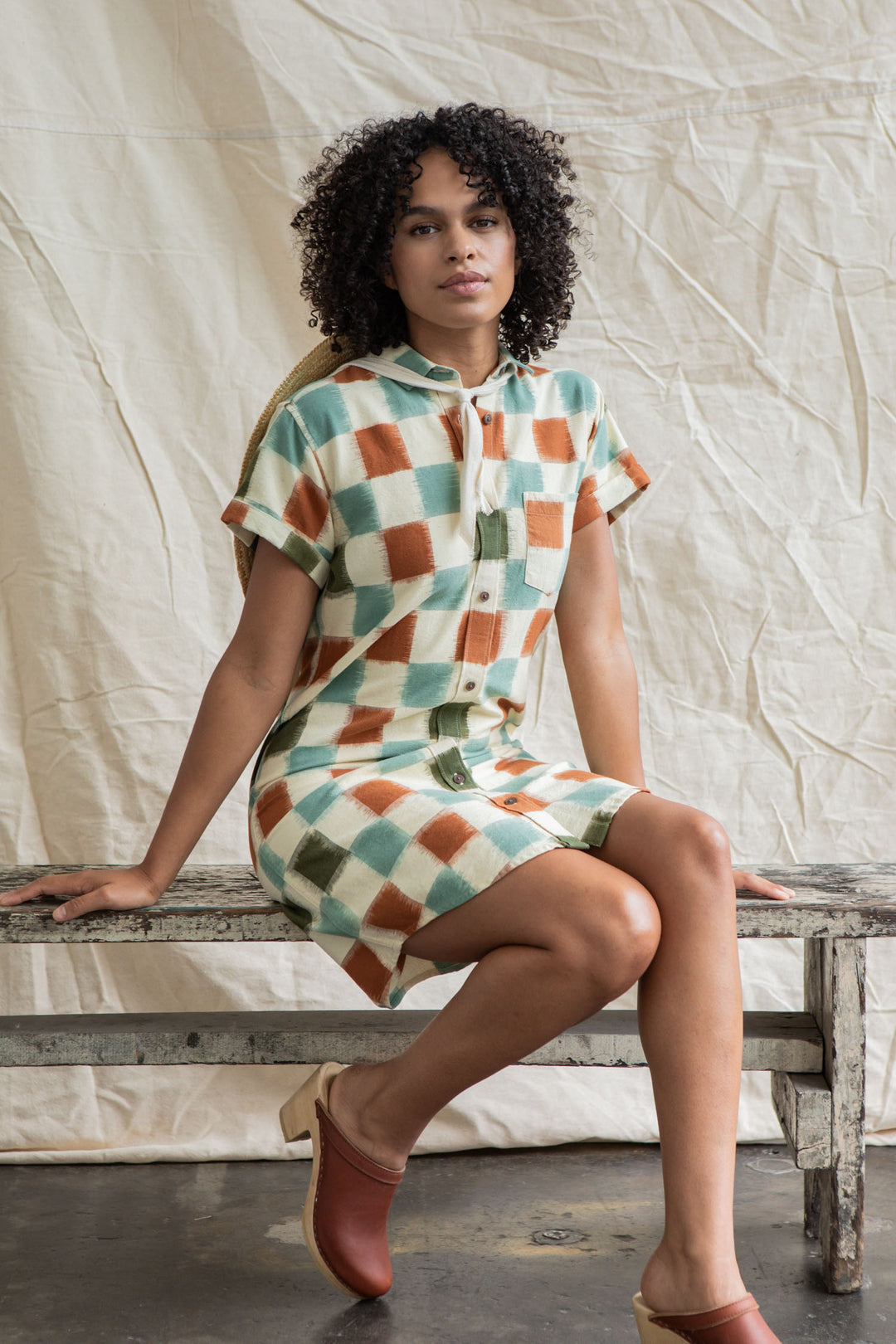 A woman with curly hair sits on a rustic bench wearing the Bridge & Burn Loren Shirt Dress in Mod Checker, paired with brown clogs. A neutral fabric backdrop hangs behind her.