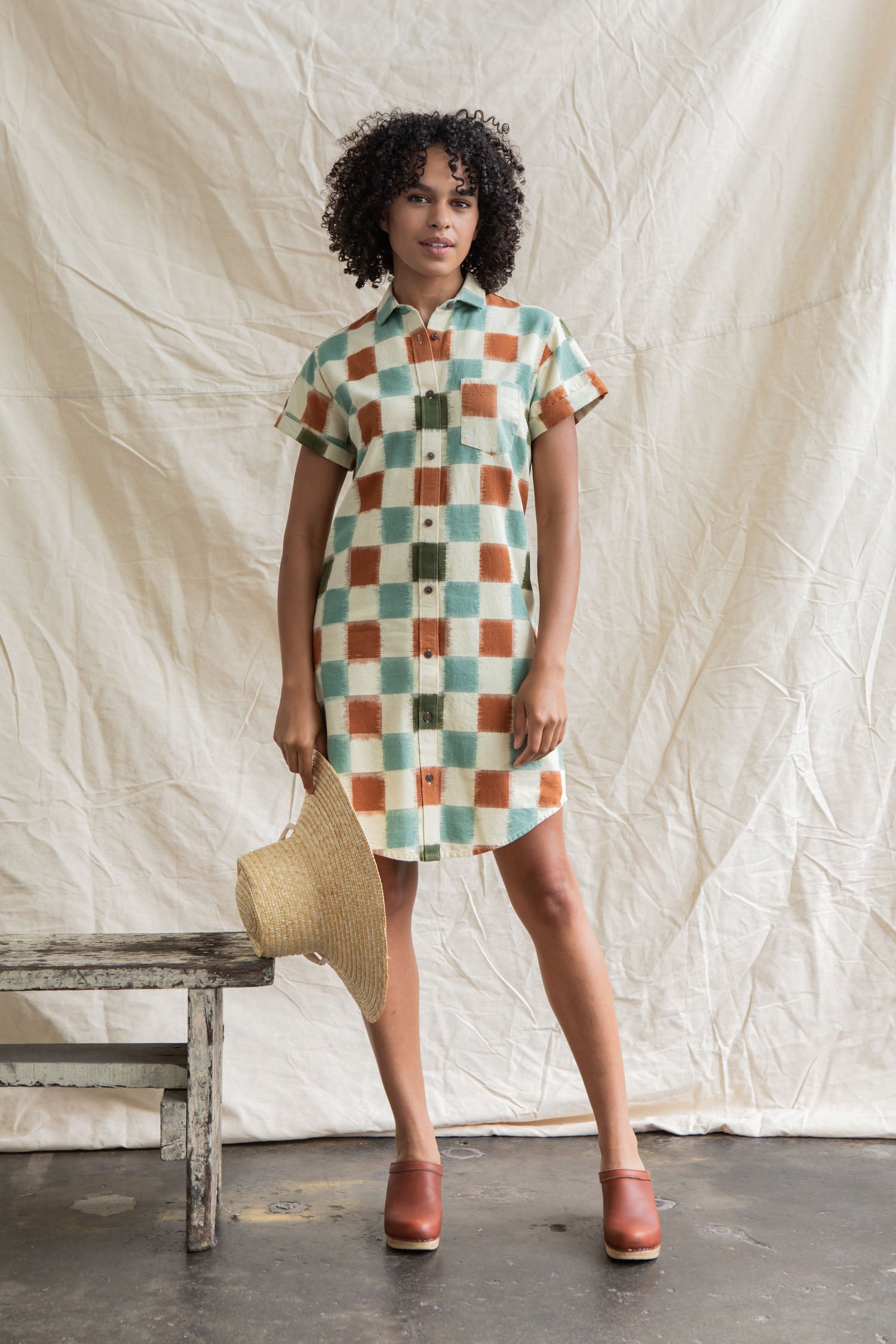 A woman with curly hair wears the Bridge & Burn Loren Shirt Dress in Mod Checker, featuring a green, orange, and white checkered 100% cotton design. She holds a straw hat beside a rustic wooden bench and beige backdrop.