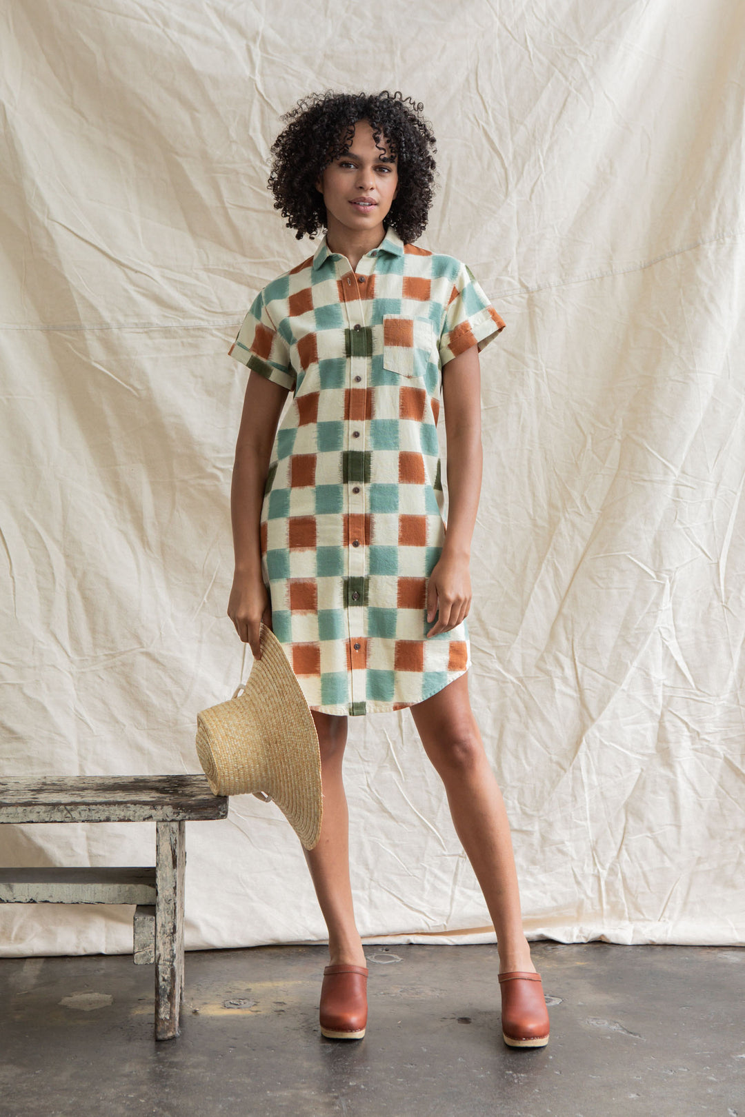 A woman with curly hair wears the Bridge & Burn Loren Shirt Dress in Mod Checker, featuring a green, orange, and white checkered 100% cotton design. She holds a straw hat beside a rustic wooden bench and beige backdrop.