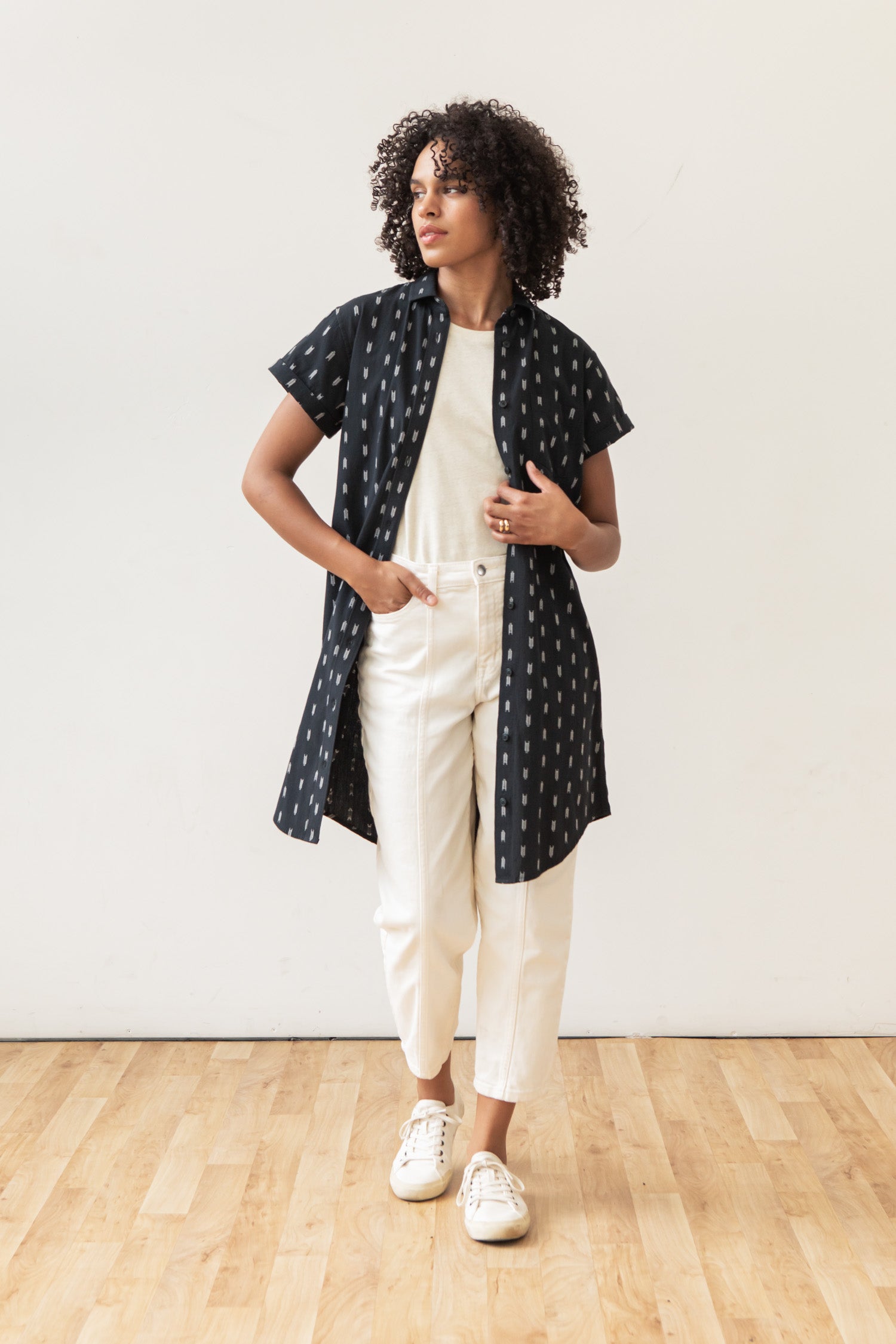A person with curly hair stands indoors on a wooden floor, wearing the Bridge & Burn Loren Shirt Dress in Onyx Arrows over a white top and pants, paired with white sneakers. The background is a plain white wall.