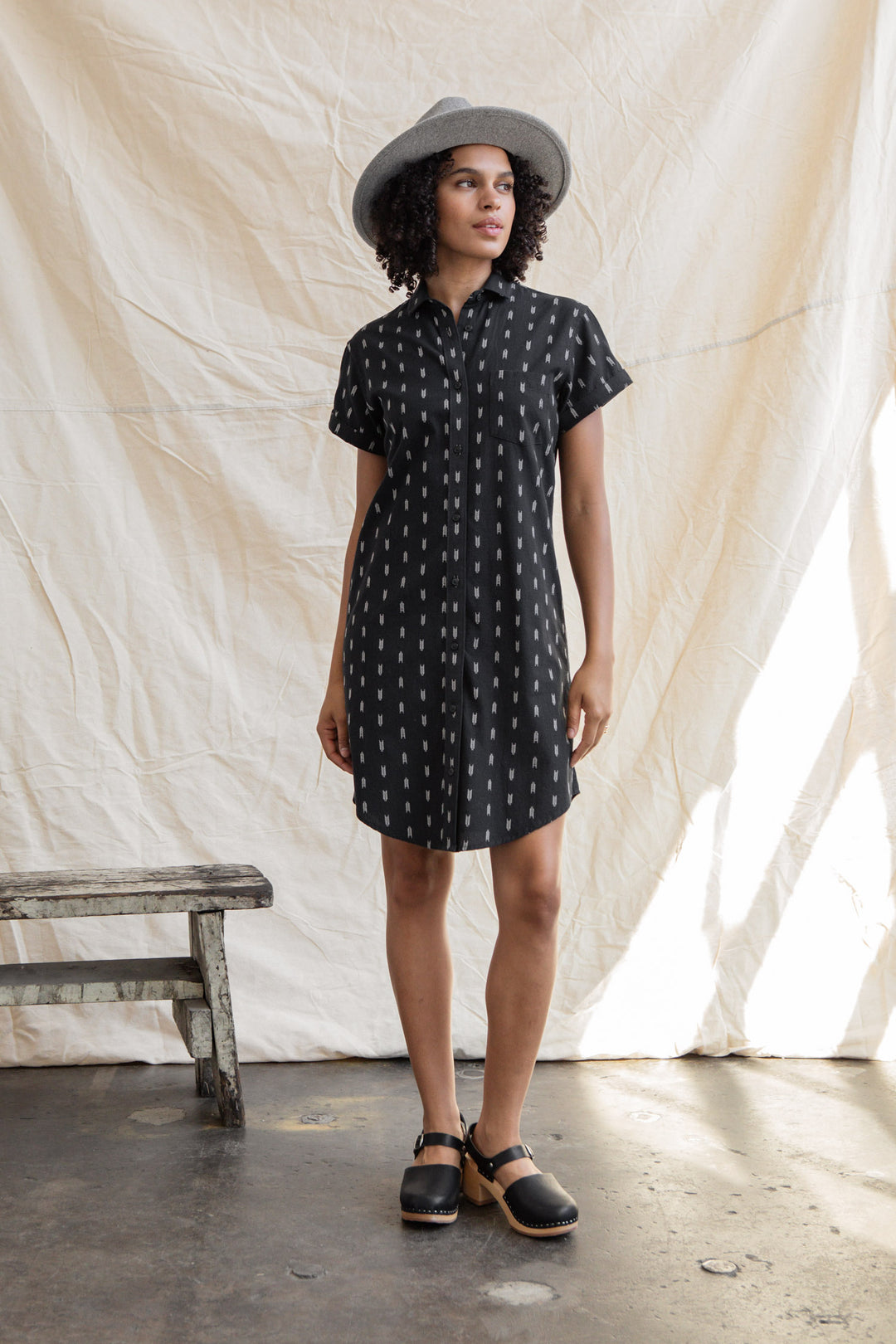 A woman stands indoors near a rustic bench, wearing the Bridge & Burn Loren Shirt Dress in Onyx Arrows, paired with a gray hat and black clogs.