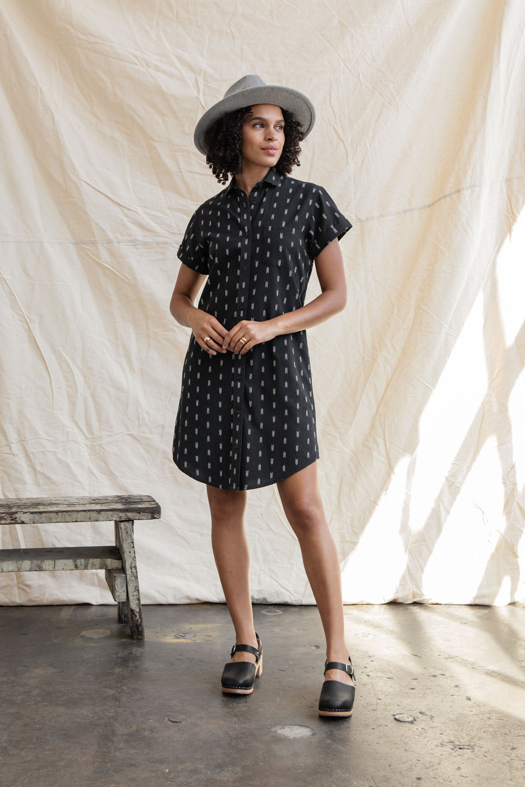 A woman stands indoors on a concrete floor near a wooden bench, wearing the Bridge & Burn Loren Shirt Dress in Onyx Arrows, a gray hat, and black clogs, with a cream-colored fabric backdrop behind her.