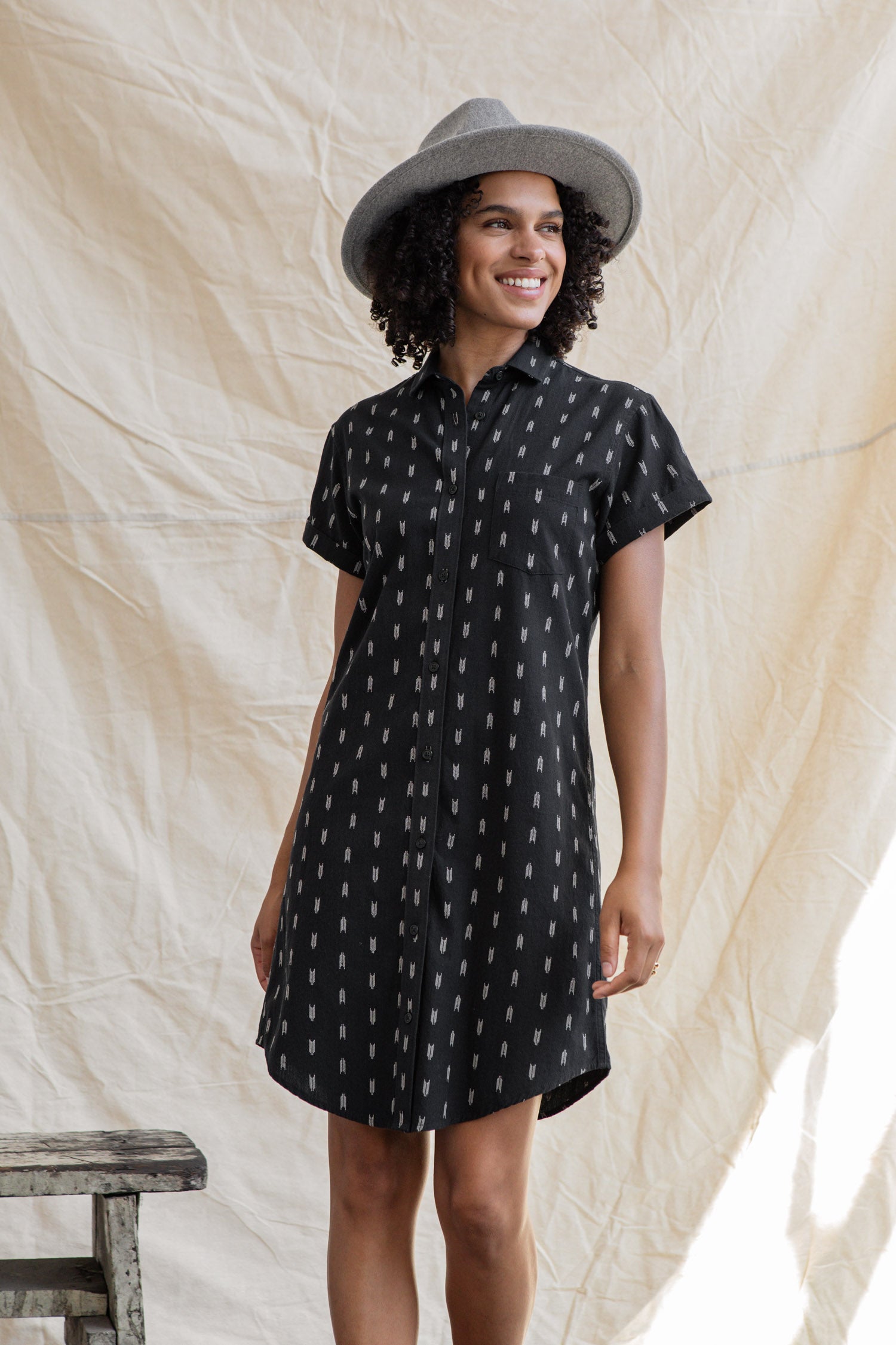A smiling woman wears the Bridge & Burn Loren Shirt Dress in Onyx Arrows—a short-sleeved, black cotton dress with a white pattern—paired with a gray hat, standing before a beige backdrop beside a rustic wooden stool.