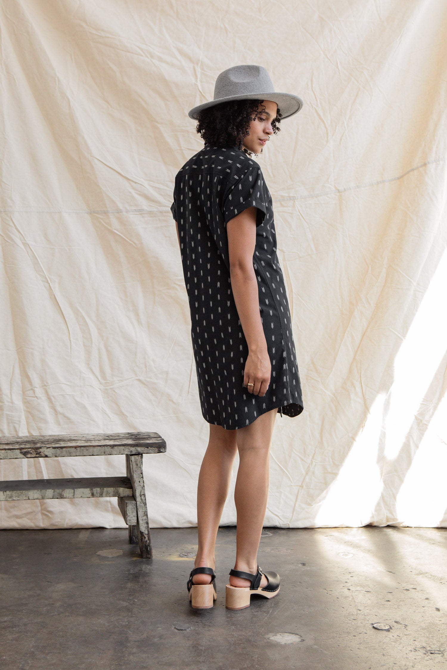 A woman in the Loren Shirt Dress / Onyx Arrows by Bridge & Burn, paired with a grey hat and black sandals, stands sideways near a rustic wooden bench before a light fabric backdrop.