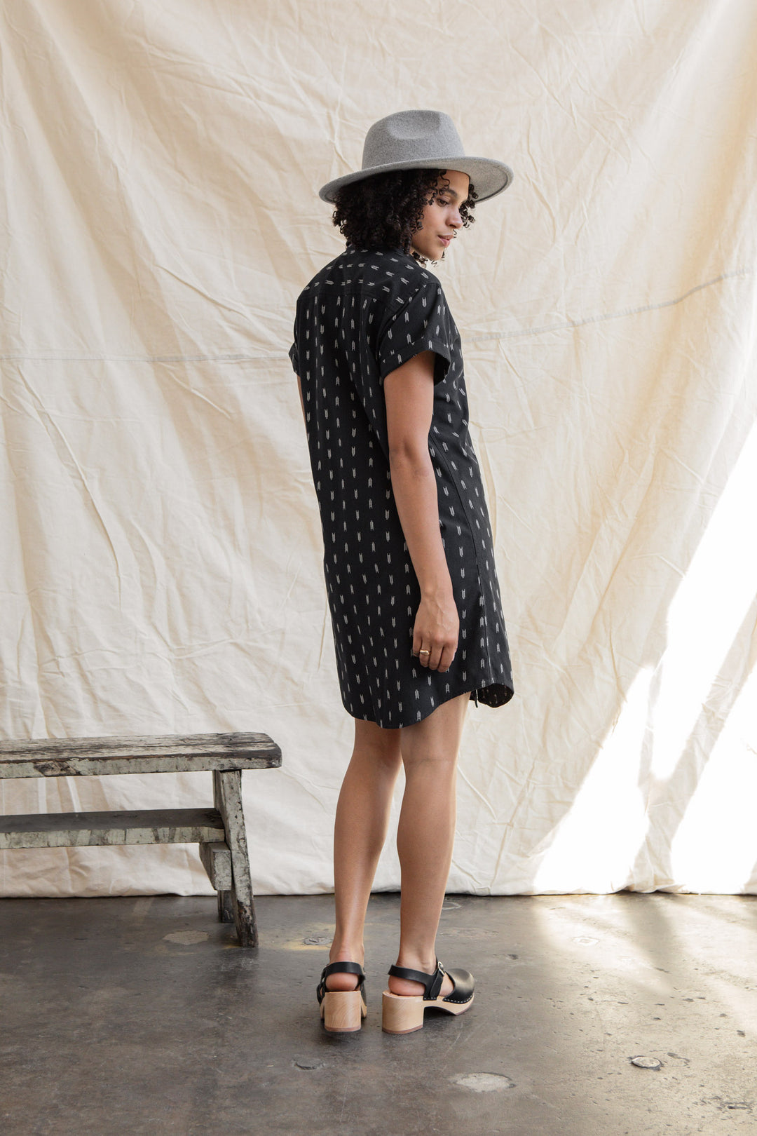 A woman in the Loren Shirt Dress / Onyx Arrows by Bridge & Burn, paired with a grey hat and black sandals, stands sideways near a rustic wooden bench before a light fabric backdrop.