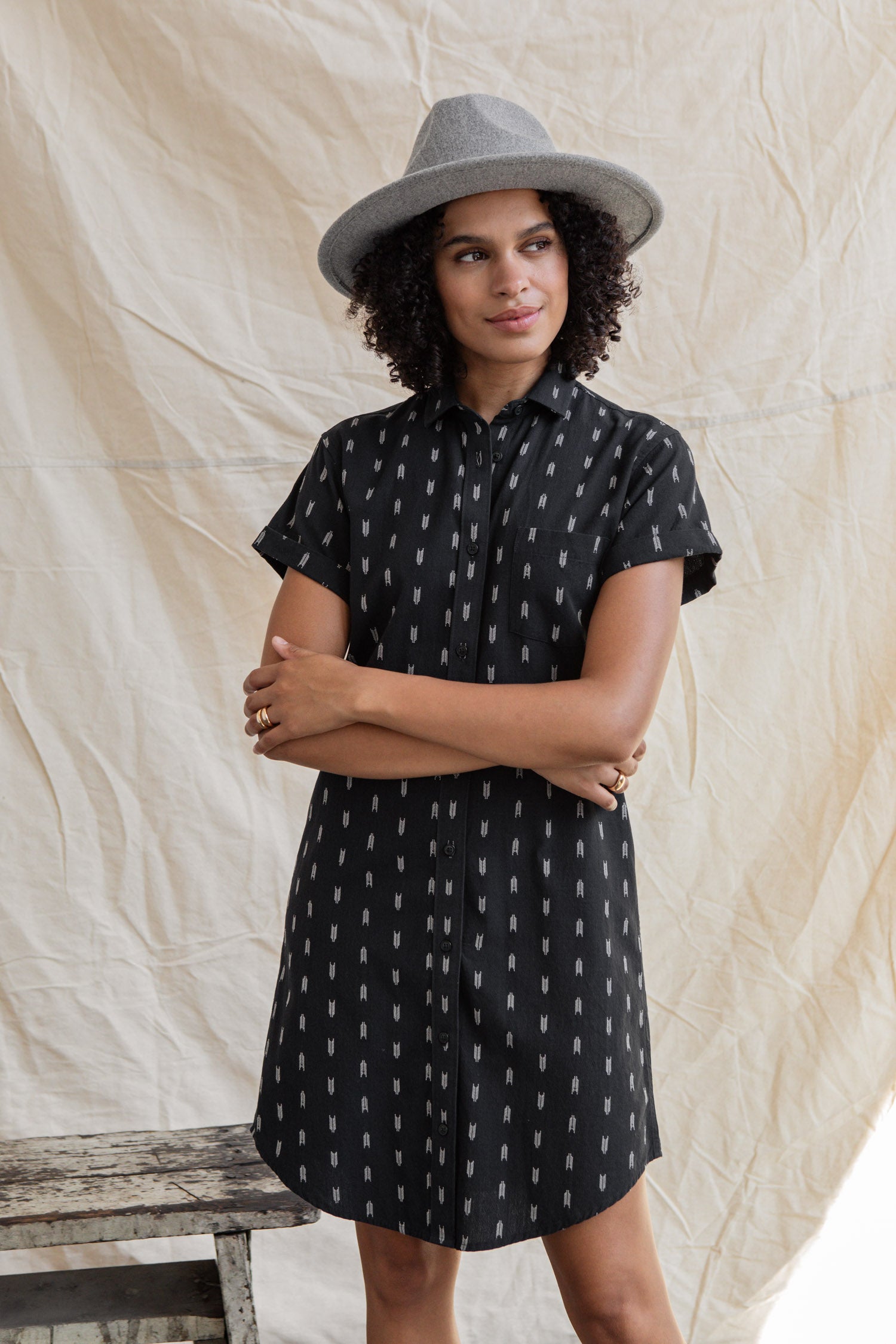 A woman with curly hair wears the Loren Shirt Dress in Onyx Arrows by Bridge & Burn, paired with a gray hat, standing with arms crossed before a light fabric backdrop.