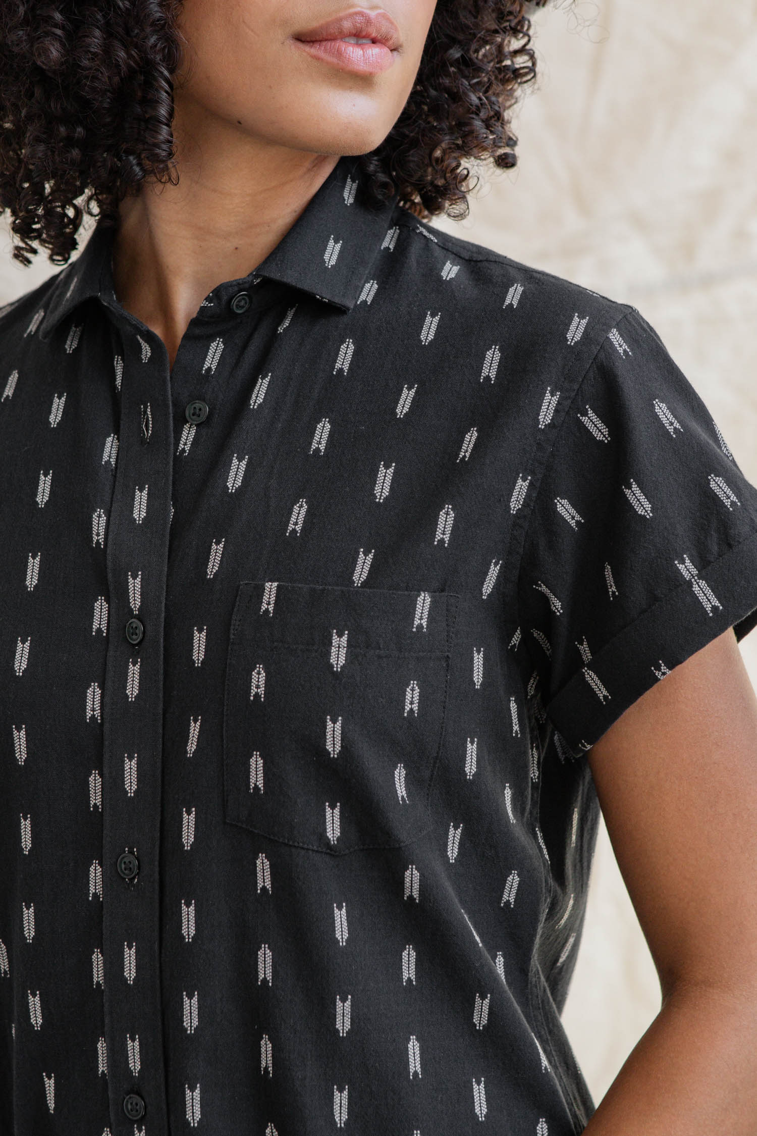 A woman with curly hair, turned slightly away from the camera, wears the Bridge & Burn Loren Shirt Dress in Onyx Arrows—a short-sleeved black cotton dress featuring a white geometric arrow pattern and a chest pocket.