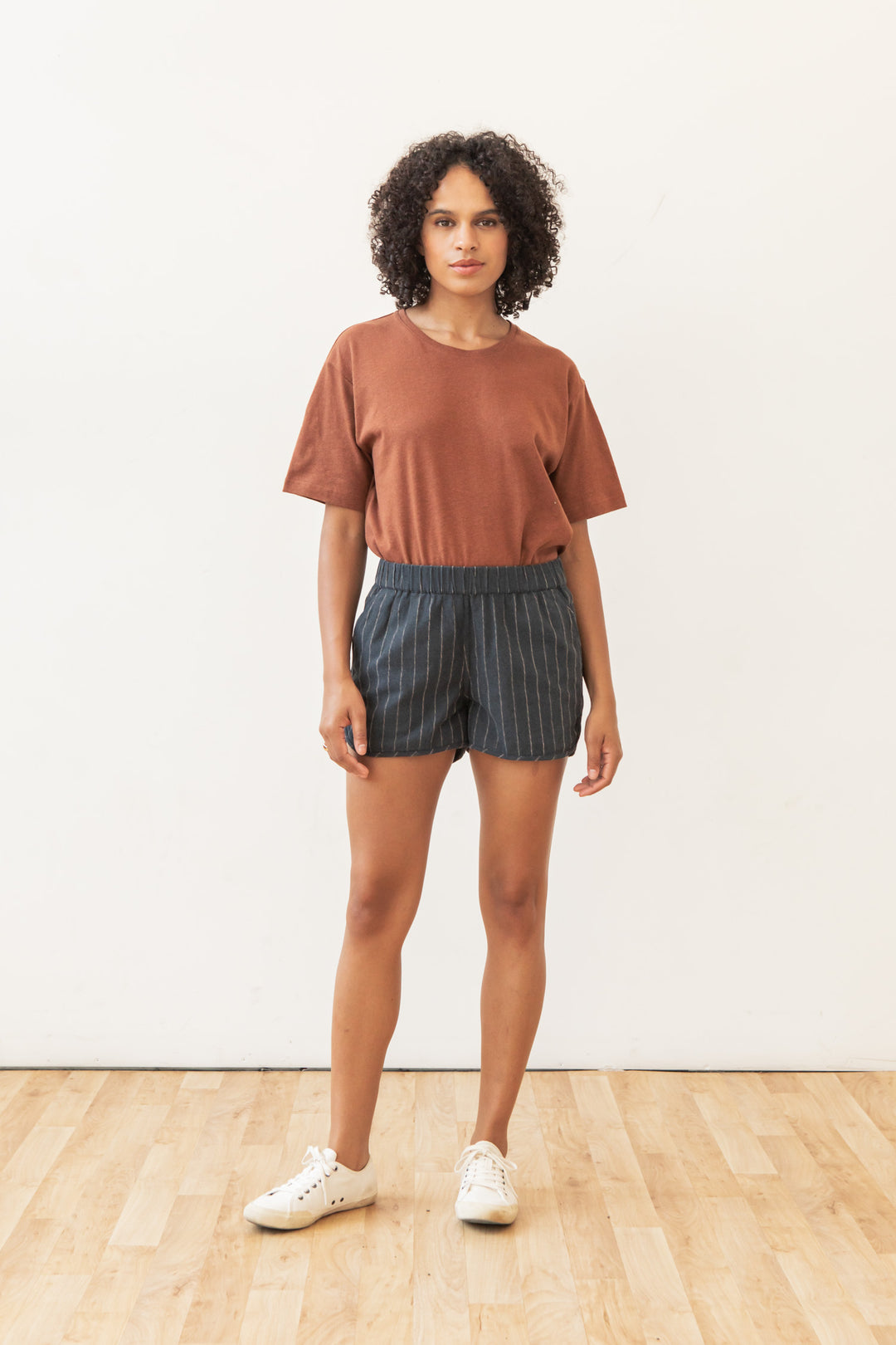 A person with curly hair stands on a wooden floor against a plain white wall, wearing a rust-colored t-shirt, Bridge & Burn Luca Track Short in Charcoal Stripe, and white sneakers.