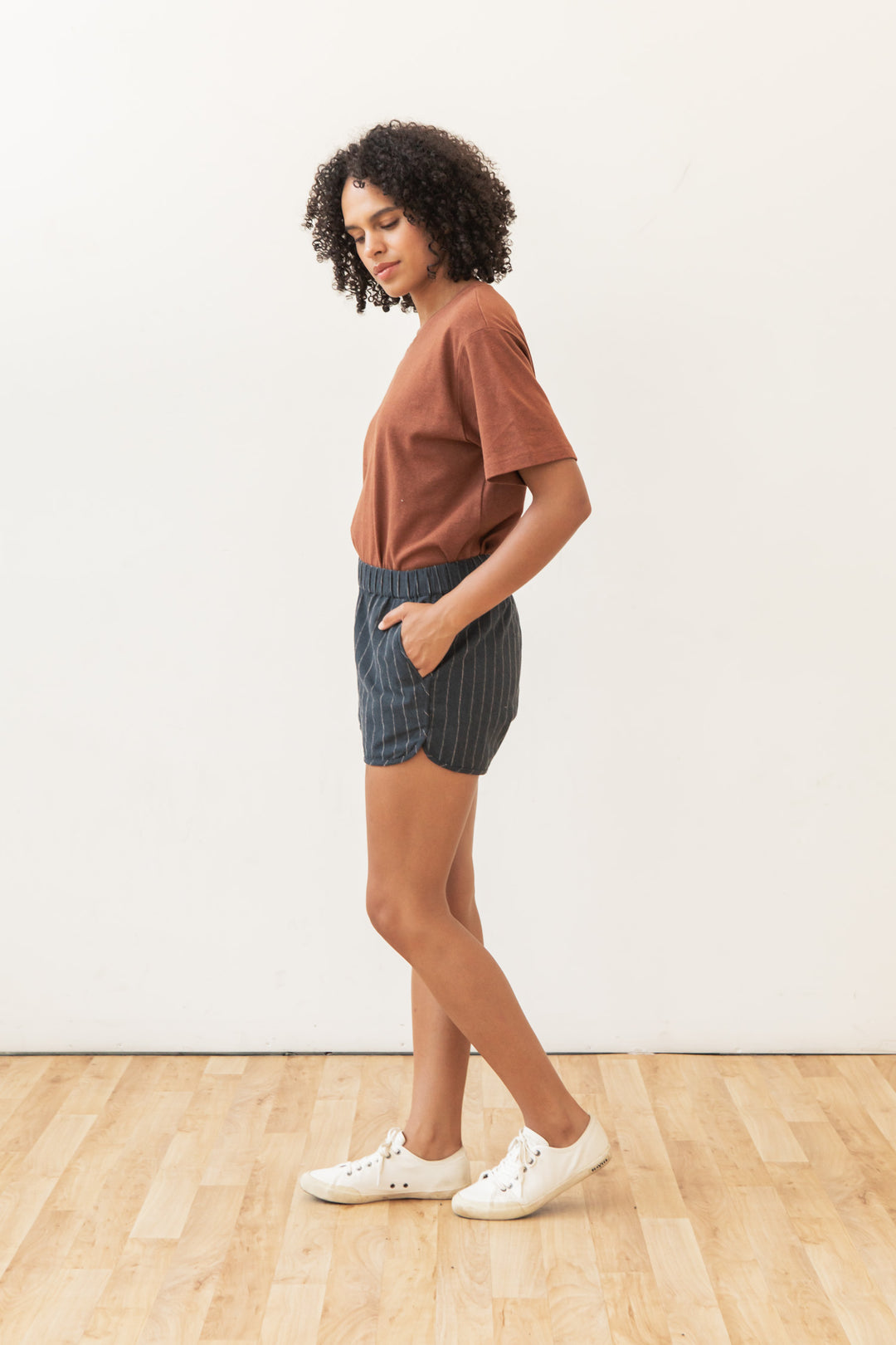 A woman with curly hair stands sideways on a wooden floor, wearing a brown t-shirt, Bridge & Burn's Luca Track Short in Charcoal Stripe, and white sneakers, with her hands in her pockets against a plain white background.
