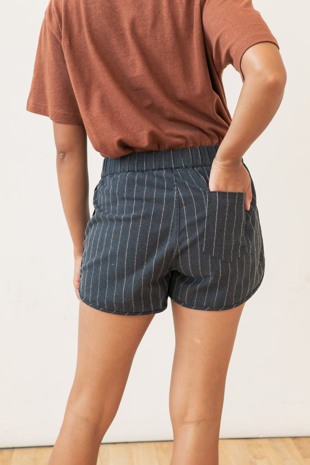 Person wearing Bridge & Burn's Luca Track Short in Charcoal Stripe—dark blue pinstriped cotton-linen summer shorts with elastic waistband and one back pocket—stands against a plain light background.