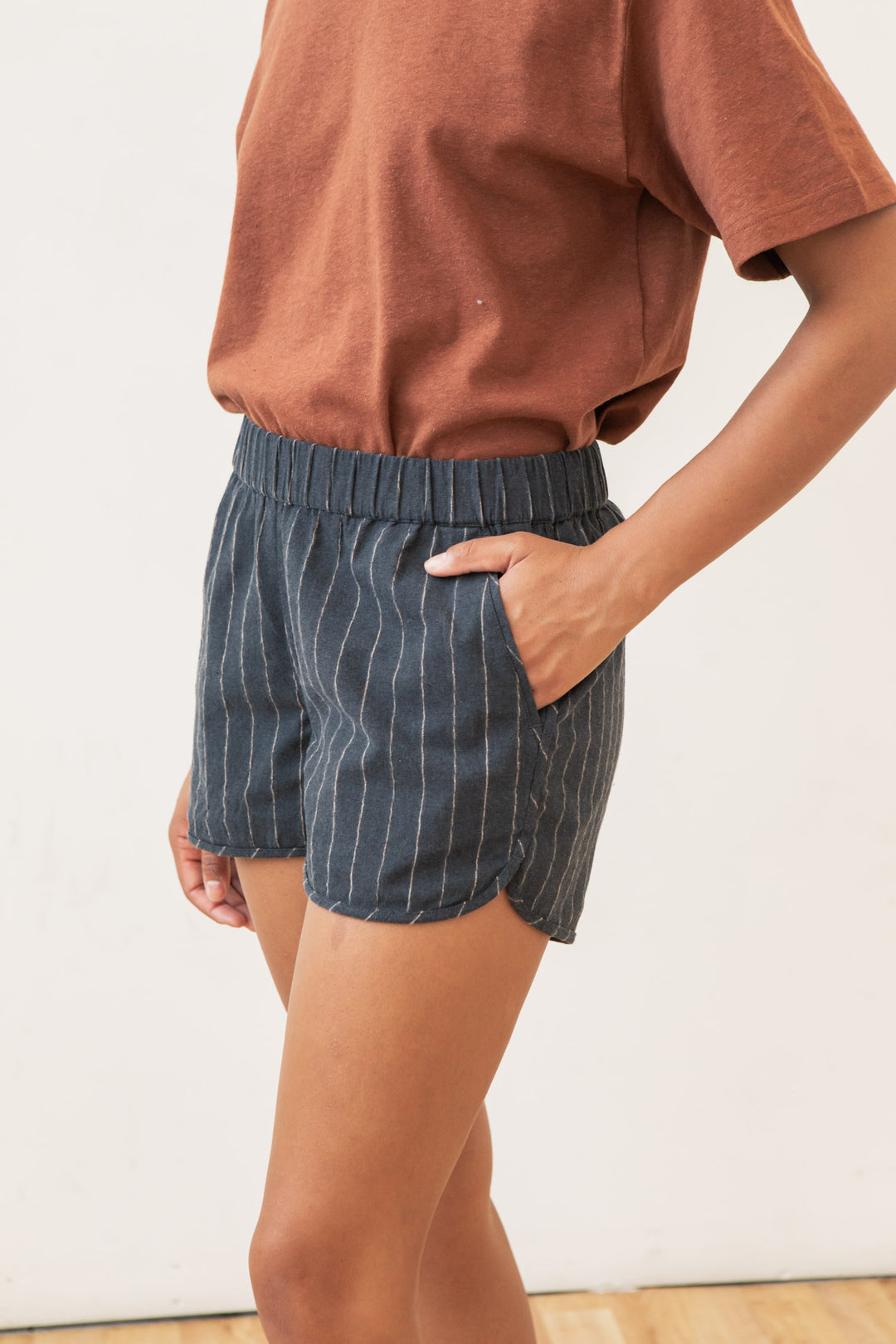 A person stands with one hand in their pocket, wearing a rust short-sleeve shirt and Bridge & Burn’s Luca Track Short in Charcoal Stripe, featuring dark blue pinstripes and an elastic waistband. The background is plain and light-colored.