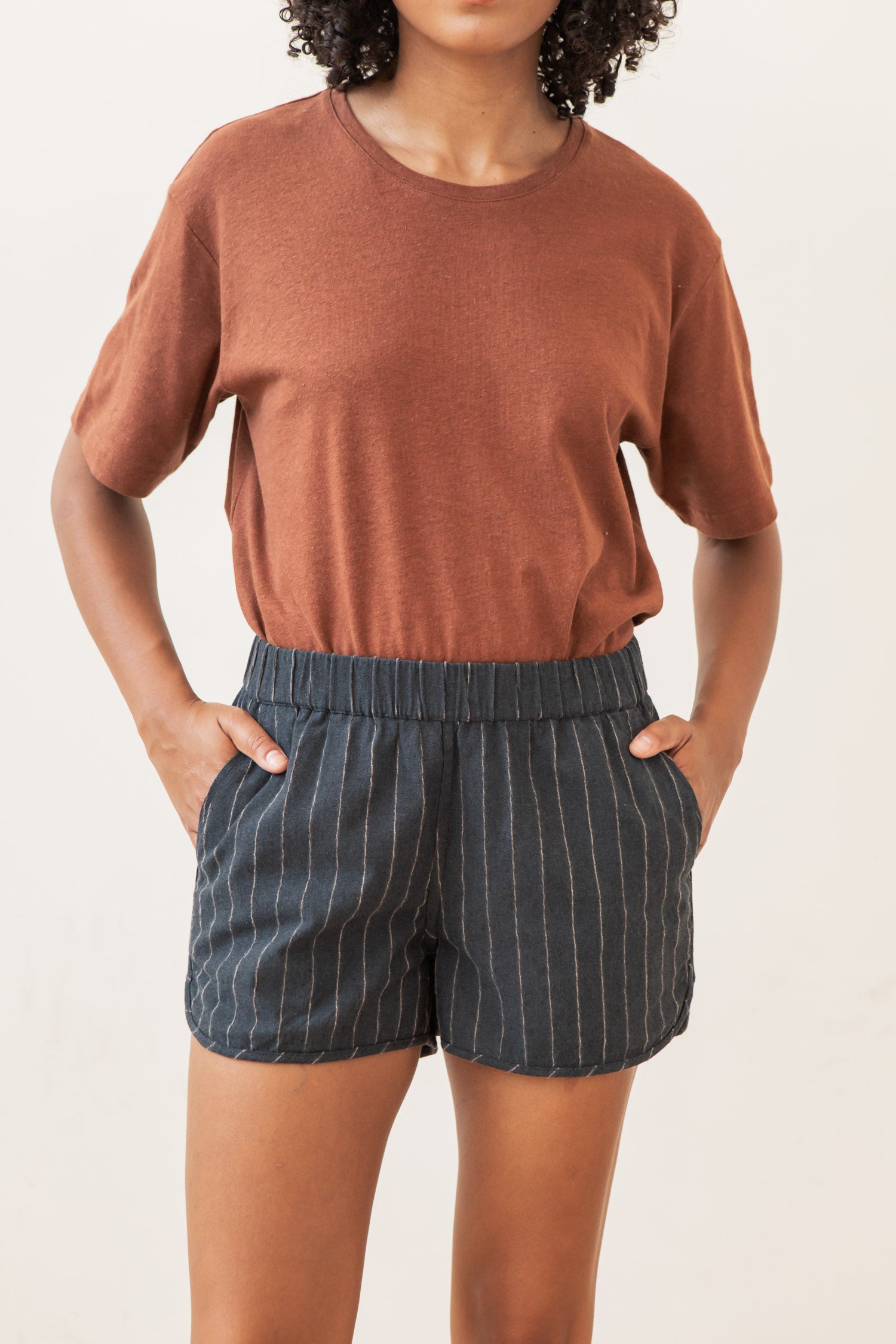 A person stands with hands in pockets, wearing Bridge & Burn’s Luca Track Short in Charcoal Stripe and a brown short-sleeve tee. The high-waisted shorts feature vertical white pinstripes. The plain, light background keeps the face out of view.