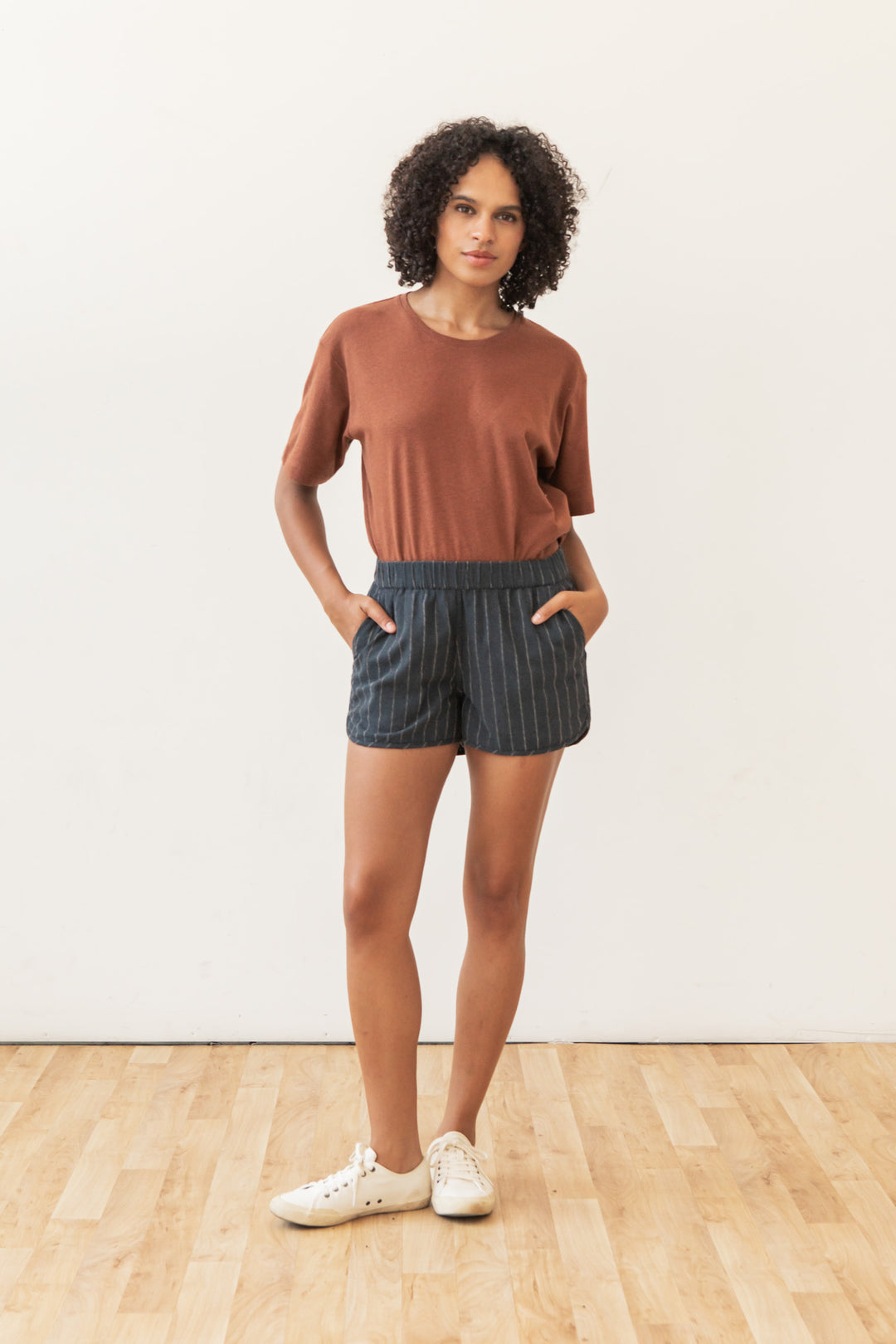 A woman with curly hair stands indoors on a wooden floor, hands in her pockets, wearing Bridge & Burn's Luca Track Short in Charcoal Stripe and a brown short-sleeve shirt, posing against a plain white wall.