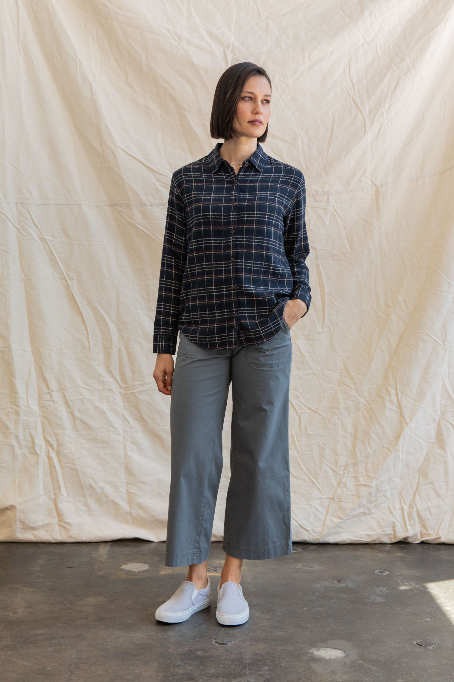 A woman stands indoors on a concrete floor, wearing the Bridge & Burn Olivia Slim Shirt in Dusk Herringbone Plaid, gray wide-leg pants, and light gray slip-on shoes. She poses with one hand in her pocket in front of a beige fabric backdrop.