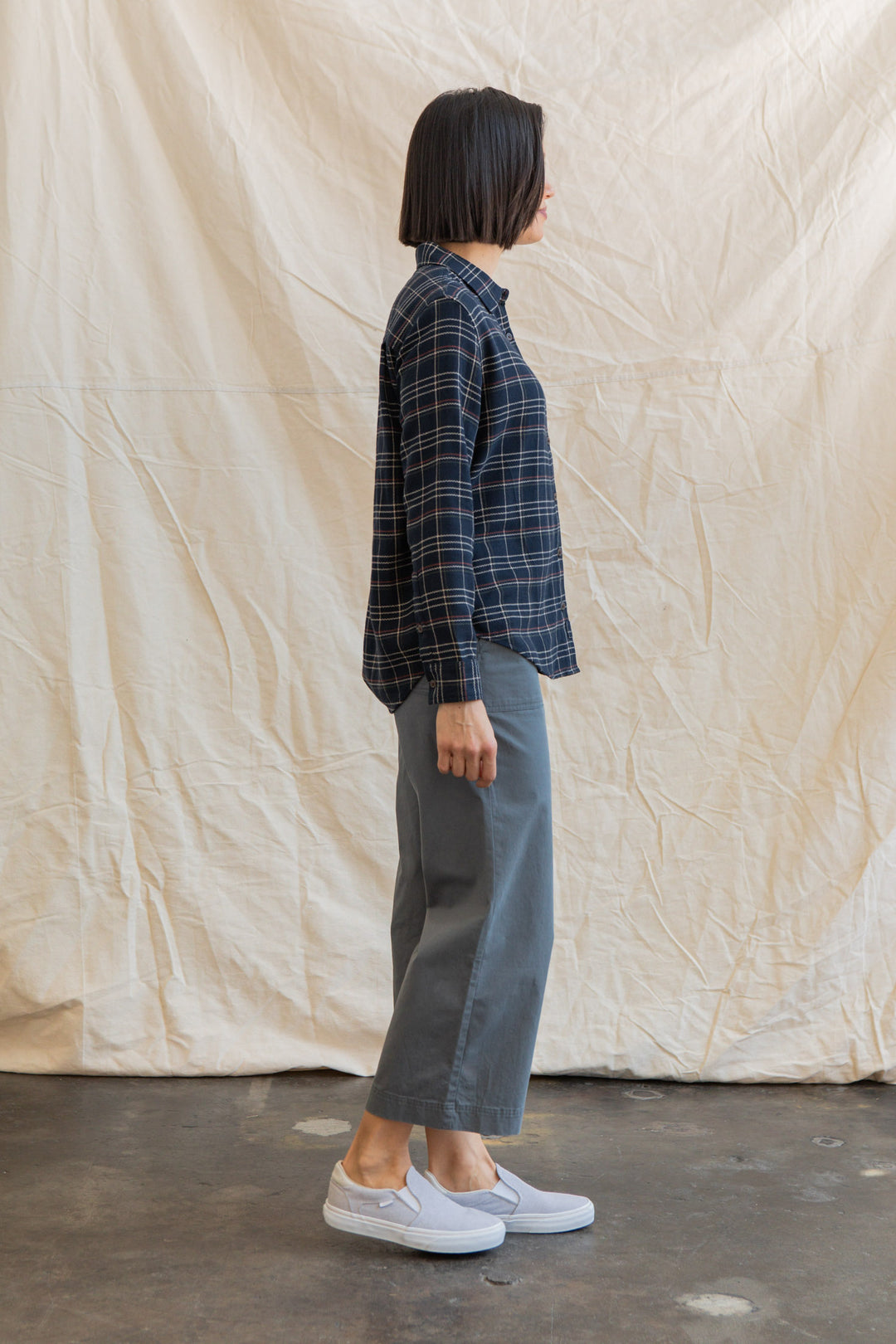 A person with straight, shoulder-length dark hair stands in profile against a cream fabric backdrop, wearing Bridge & Burn’s Olivia Slim Shirt in Dusk Herringbone Plaid, gray wide-leg pants, and light gray slip-on shoes.