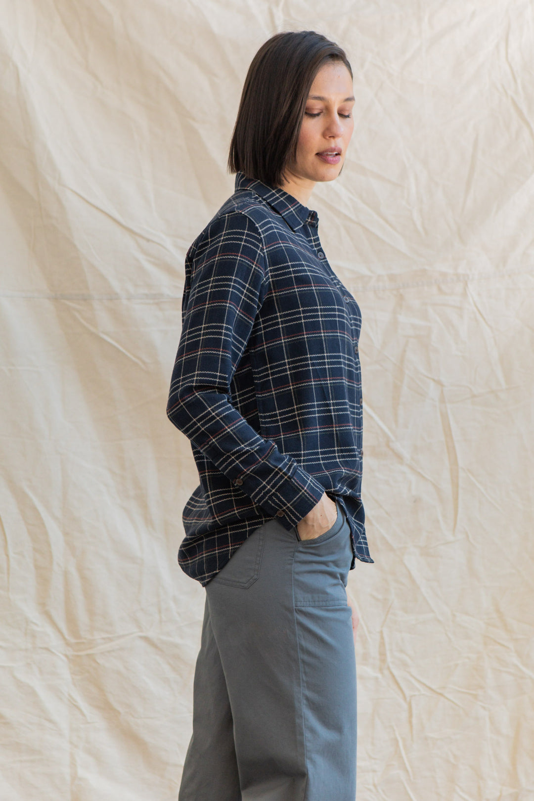 A person with straight, shoulder-length dark hair stands sideways, hand in pocket, wearing the Bridge & Burn Olivia Slim Shirt in Dusk Herringbone Plaid with gray pants, posed before a light beige, wrinkled fabric backdrop.