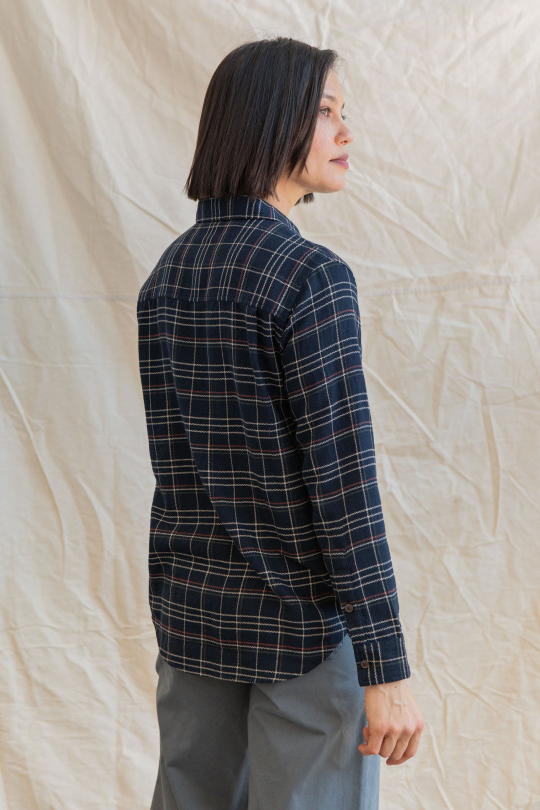 A person with short dark hair, seen from the back and slightly turned to the side, wears the Bridge & Burn Olivia Slim Shirt in Dusk Herringbone Plaid and gray pants, standing before a light, wrinkled fabric backdrop.
