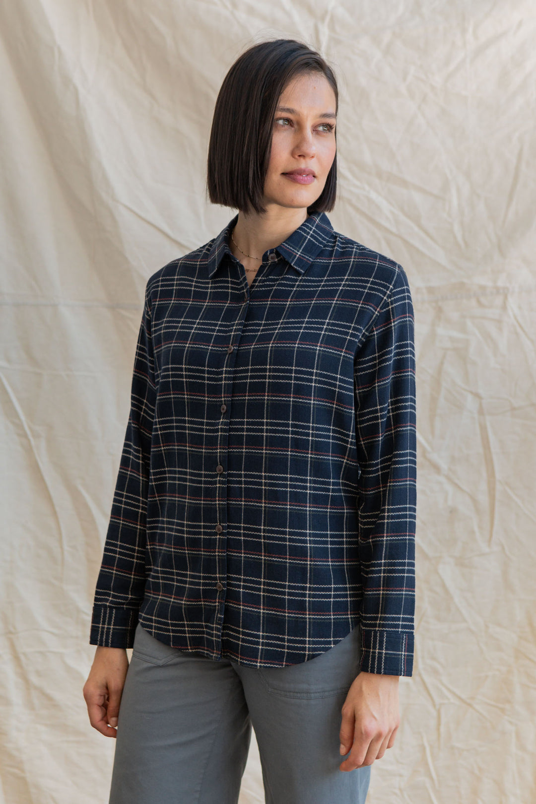 A woman with straight shoulder-length dark hair wears the Bridge & Burn Olivia Slim Shirt in Dusk Herringbone Plaid and gray pants, standing in front of a light-colored fabric backdrop.