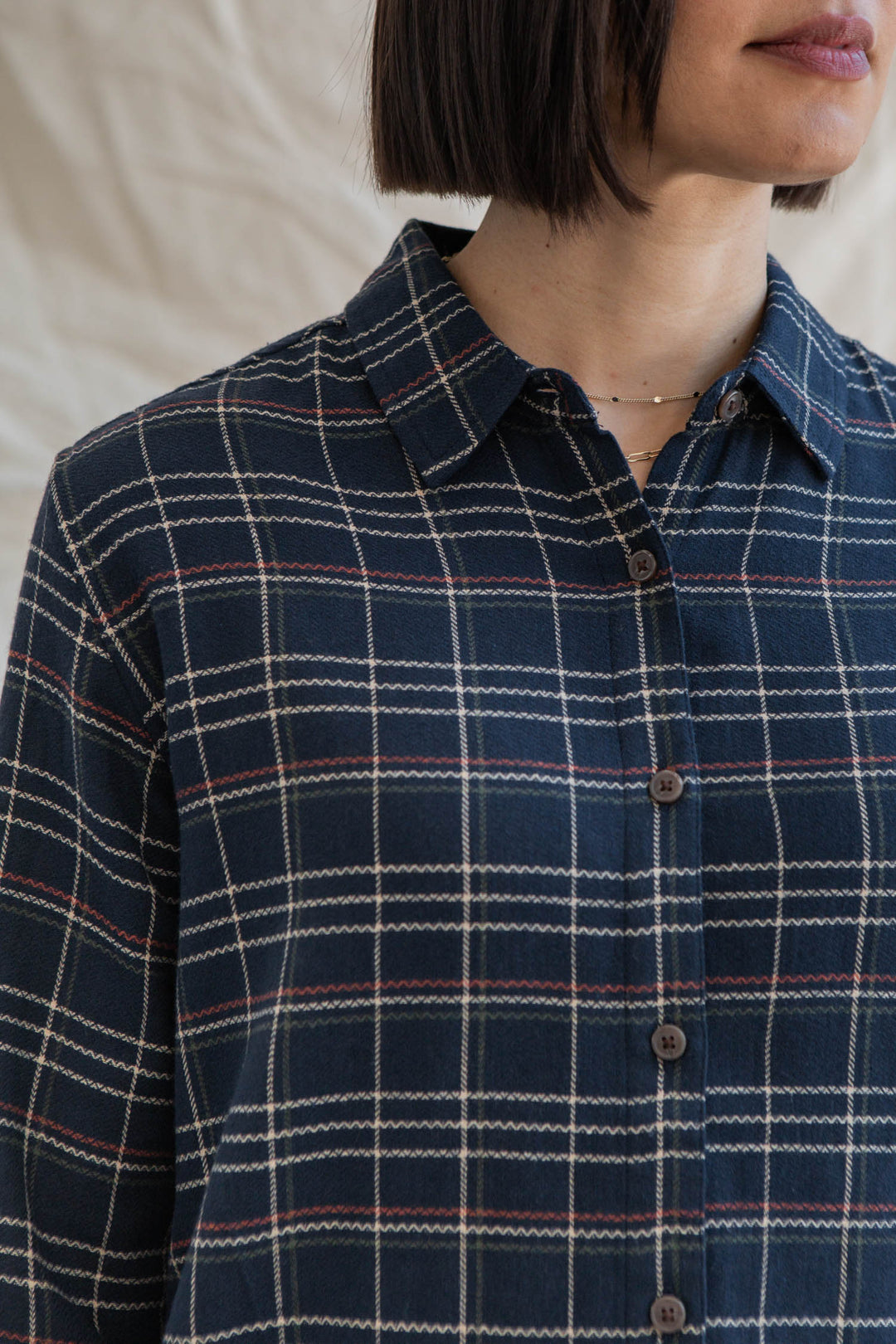 A person with short dark hair models the Bridge & Burn Olivia Slim Shirt in Dusk Herringbone Plaid, styled with a gold necklace. The image is cropped at the chin and mid-torso.