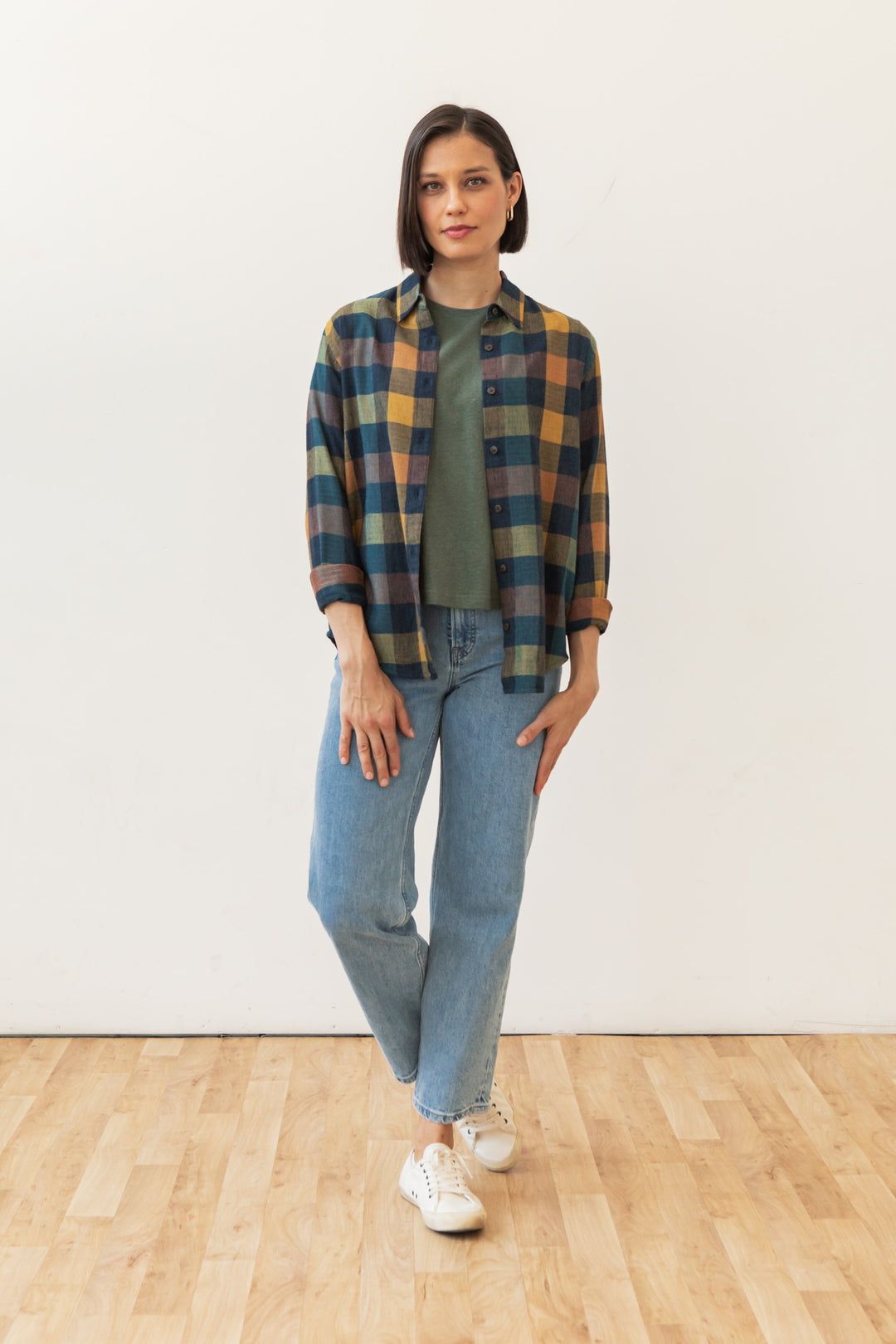 A woman with short brown hair stands indoors on a wooden floor, wearing the Bridge & Burn Olivia Slim Shirt in Moonbow Plaid over a green top, light blue jeans, and white sneakers, her expression calm and relaxed.