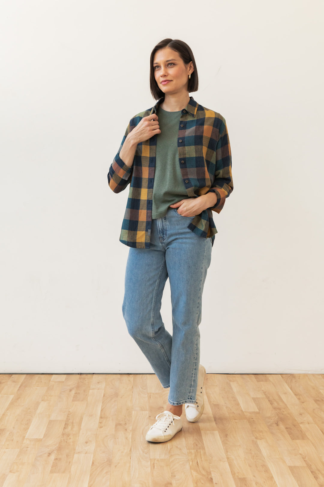 A woman stands indoors on a wooden floor against a white wall, wearing the Bridge & Burn Olivia Slim Shirt in Moonbow Plaid, light blue jeans, and white sneakers. One hand is in her pocket and the other touches her shirt collar.
