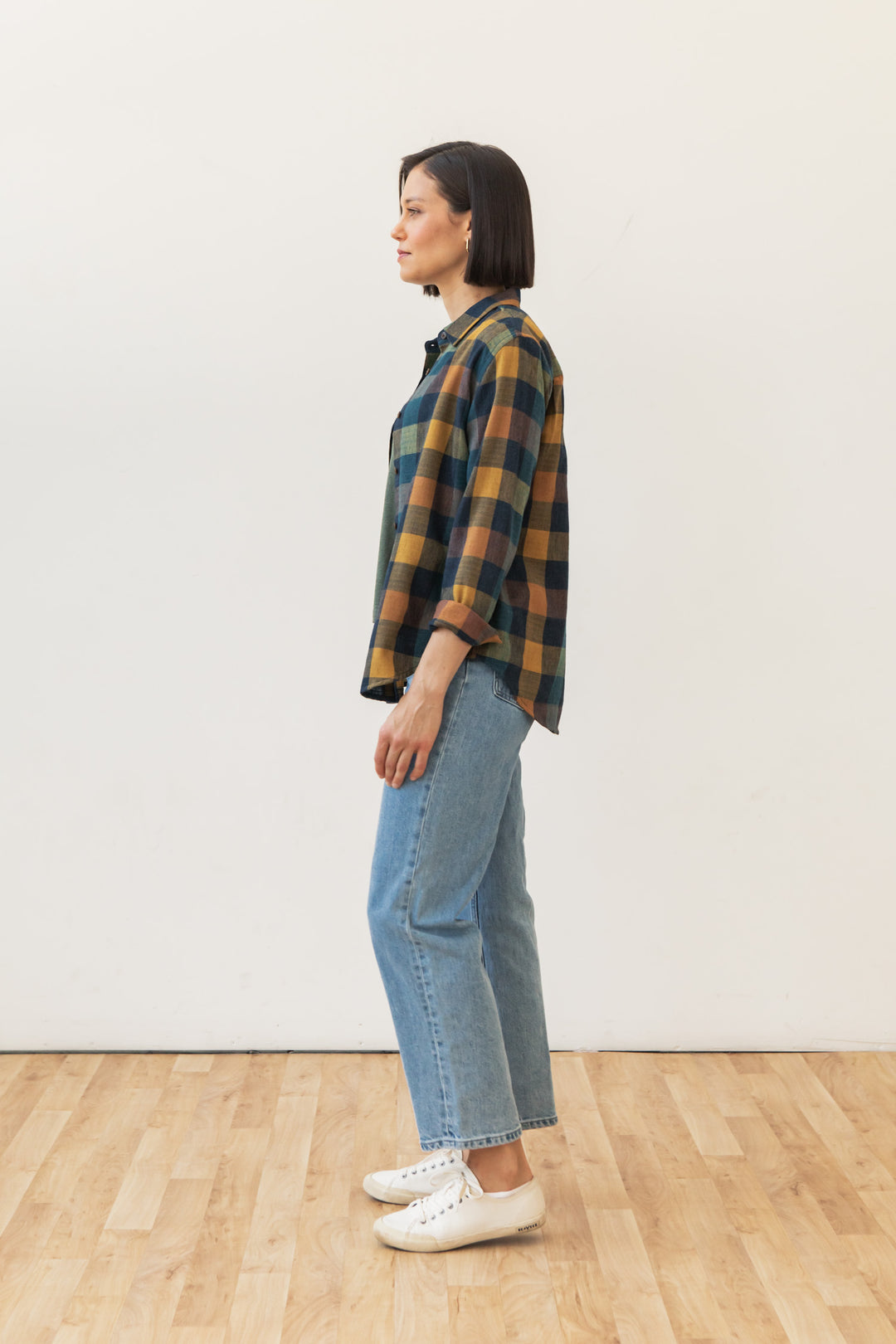 A person with short dark hair stands in profile on a wooden floor, wearing the Bridge & Burn Olivia Slim Shirt in Moonbow Plaid, light blue jeans, and white shoes, against a plain off-white wall.