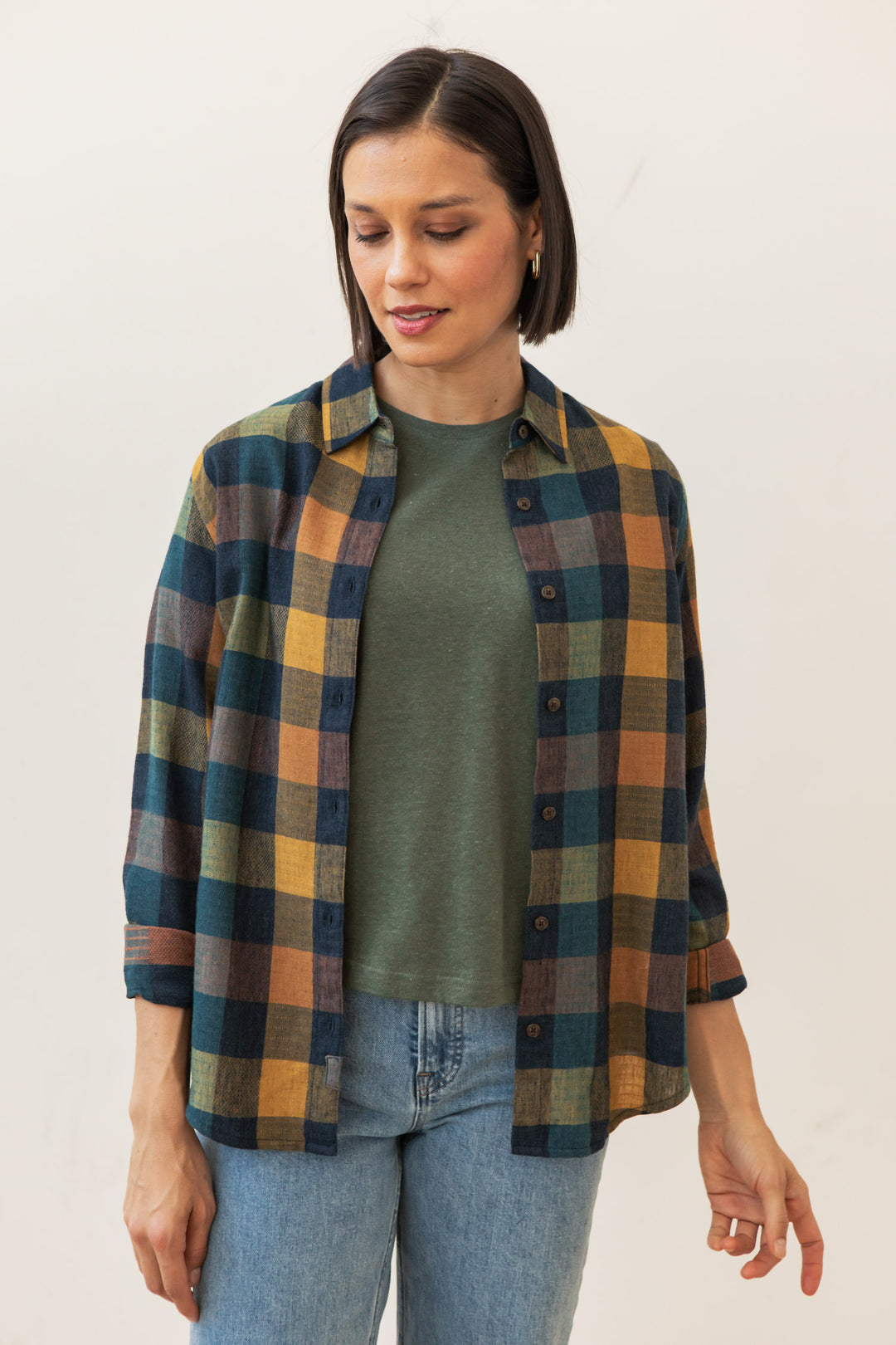 A woman with short dark hair wears a green tee, light blue jeans, and the Bridge & Burn Olivia Slim Shirt in Moonbow Plaid. She looks down with a slight smile against a plain light background.