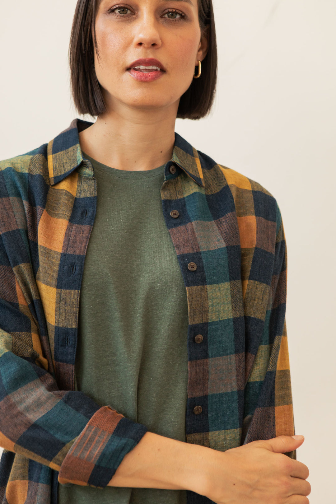 A woman with short dark hair wears the Bridge & Burn Olivia Slim Shirt in Moonbow Plaid over a green tee, standing against a light background and looking at the camera with a neutral expression.