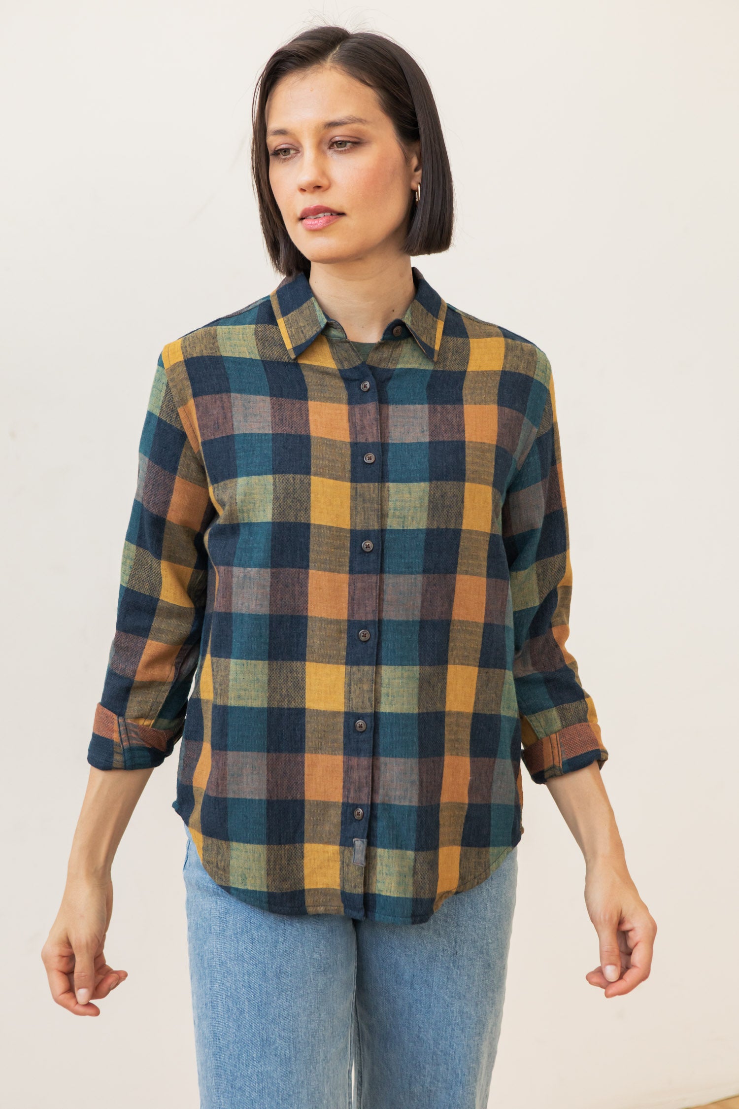 A woman with a bob wears the Bridge & Burn Olivia Slim Shirt in Moonbow Plaid—blue, yellow, and green recycled cotton—paired with light blue jeans, standing against a plain light background.