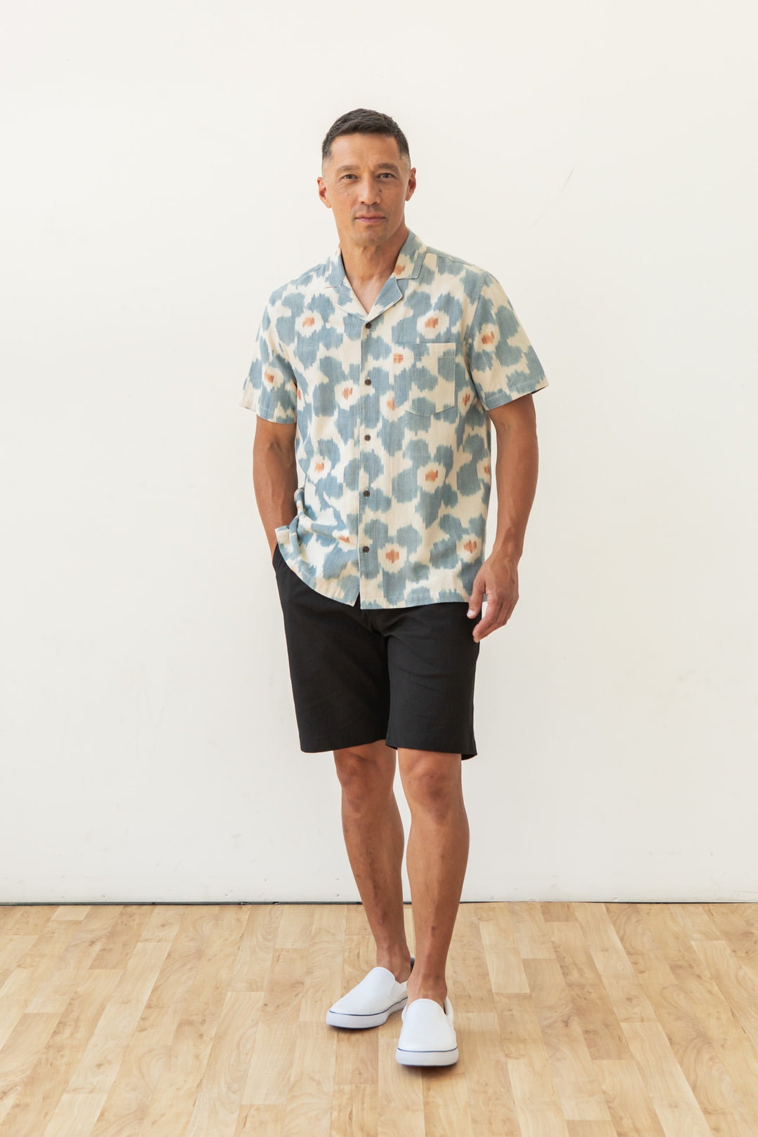 A man stands indoors on a wooden floor in front of a white wall, wearing the Bridge & Burn Bodie Shirt in Polar Blossom, black shorts, and white slip-on shoes, with one hand in his pocket.